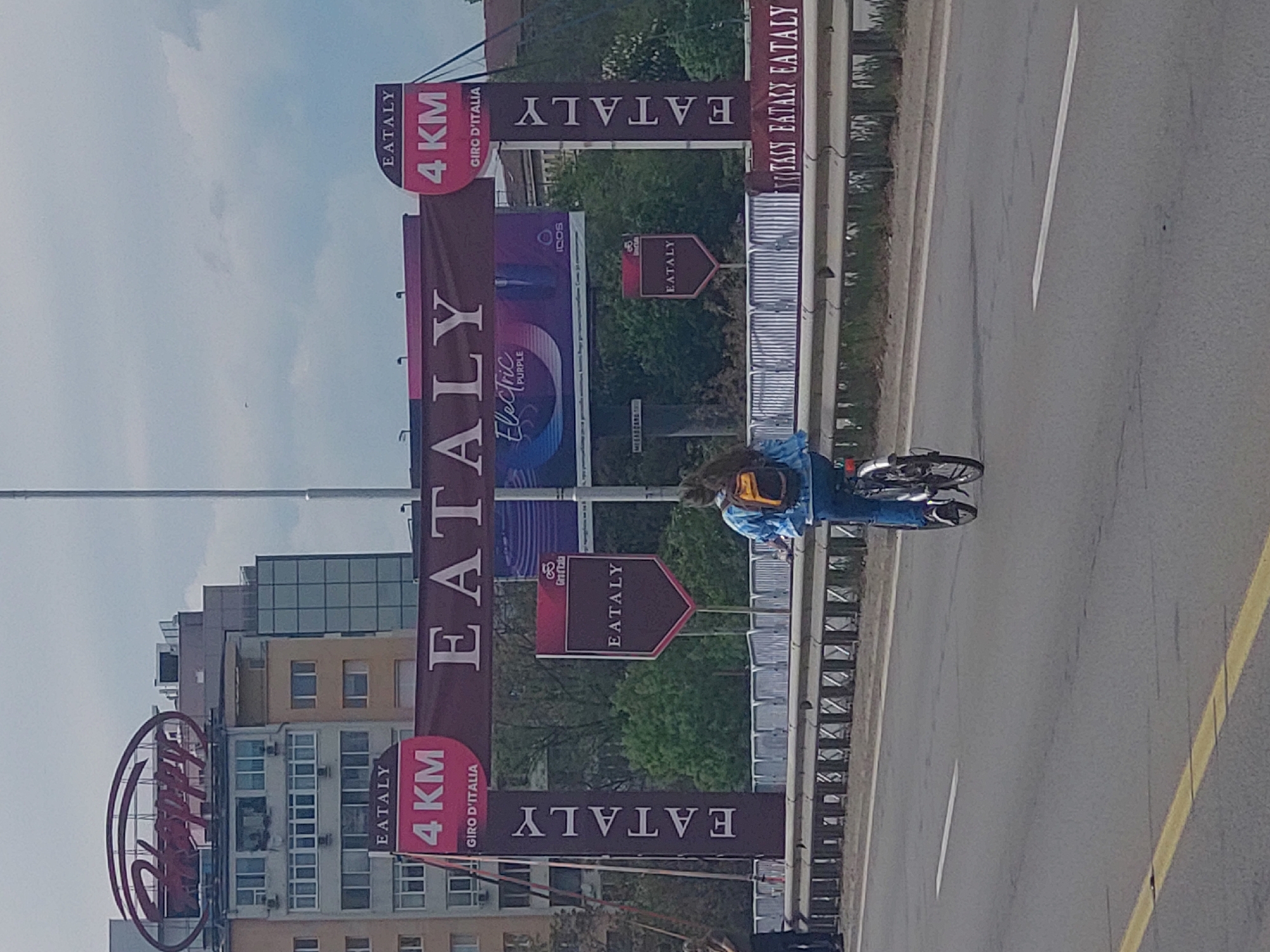 The image shows a street scene in Sofia, Bulgaria, featuring promotional banners for Eataly and the Giro d'Italia. The banners, displaying the Eataly logo and the text "4 KM Giro d'Italia," are positioned along the roadside. A cyclist, wearing blue jeans and a backpack, rides along the road next to the banners. Behind the banners are various buildings, including a structure with the "Eataly" logo. The sky is overcast. The road is marked with a yellow line.
