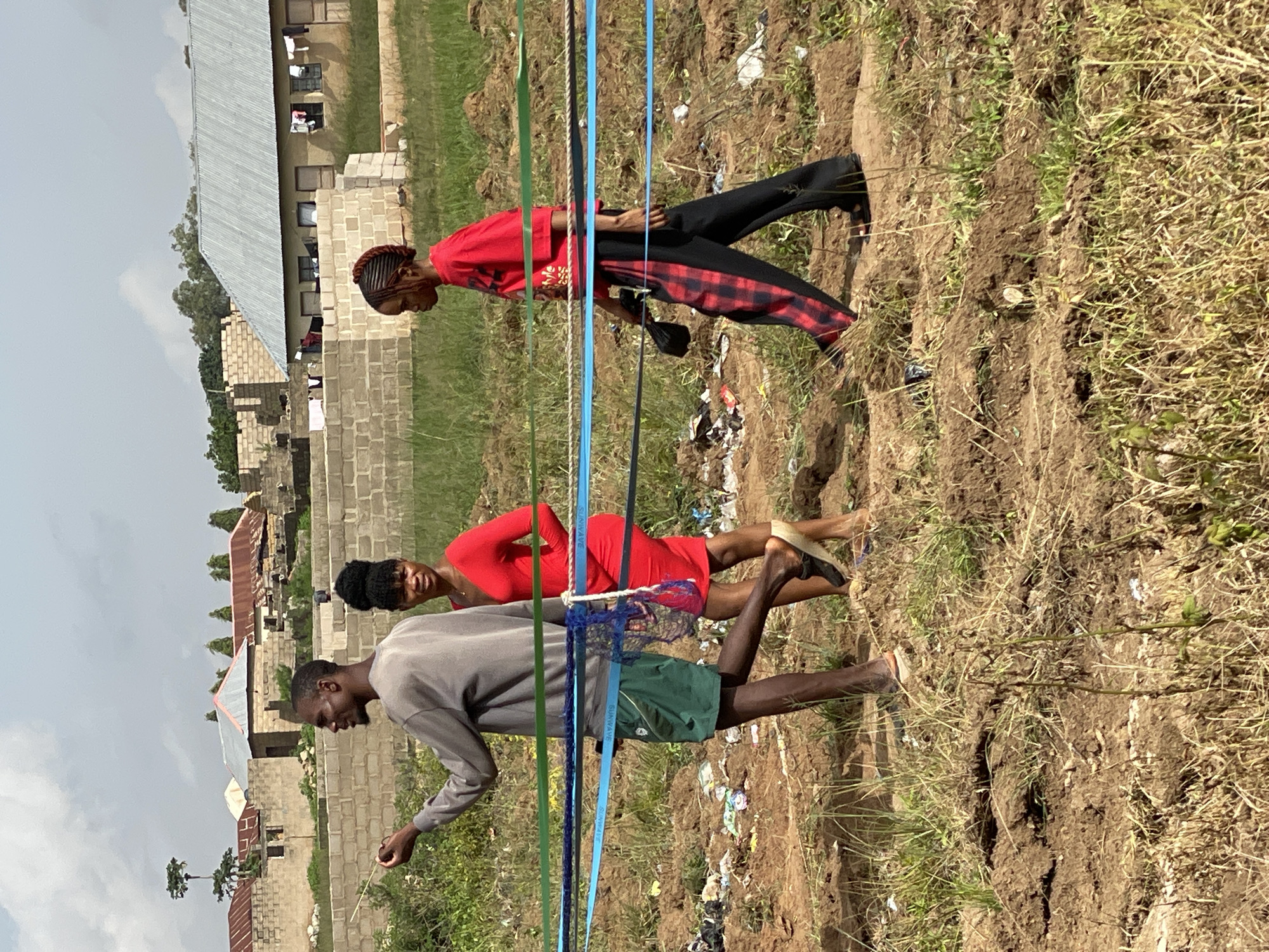 This media file shows three young adults in an outdoor setting in Durimi, Nigeria. The scene appears to be a field with dry grass and some tilled earth, possibly a construction site or farmland. In the background, there are buildings with corrugated metal roofs and some unfinished brick structures. The sky is overcast.
In the foreground, a young man in a grey long-sleeved shirt and green shorts is leaning forward, holding a thin stick. Beside him, a young woman in a red dress is standing, looking towards the camera. Further back, another young woman in a red top and black and red checkered pants is standing, also looking away from the camera. Several colorful ribbons or ropes are strung horizontally across the frame between the people.
The time of day appears to be daytime, given the natural light. The weather seems to be warm and dry. The overall atmosphere is casual, and the people do not display strong emotions. There is some litter visible in the tilled soil. The visible text on the blue ribbon reads "SUNWAYE".