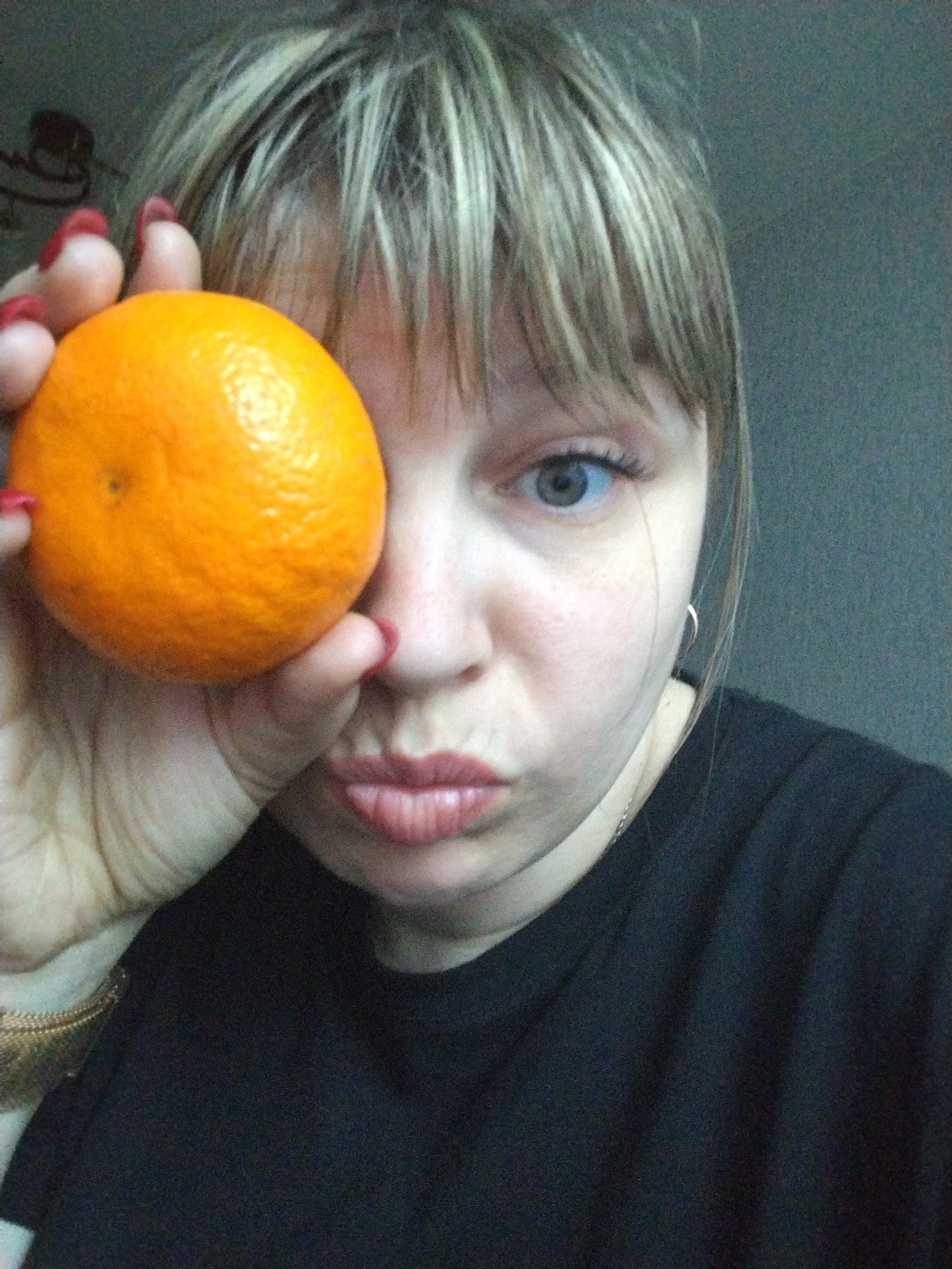 A fair-skinned woman with blonde hair and blue eyes holds an orange up to her face, covering one eye, and makes a kissing face. She is wearing a black t-shirt and a gold bracelet. The background is a gray wall. There is no information provided to suggest a location in the city of Yaroslavl, Russia.