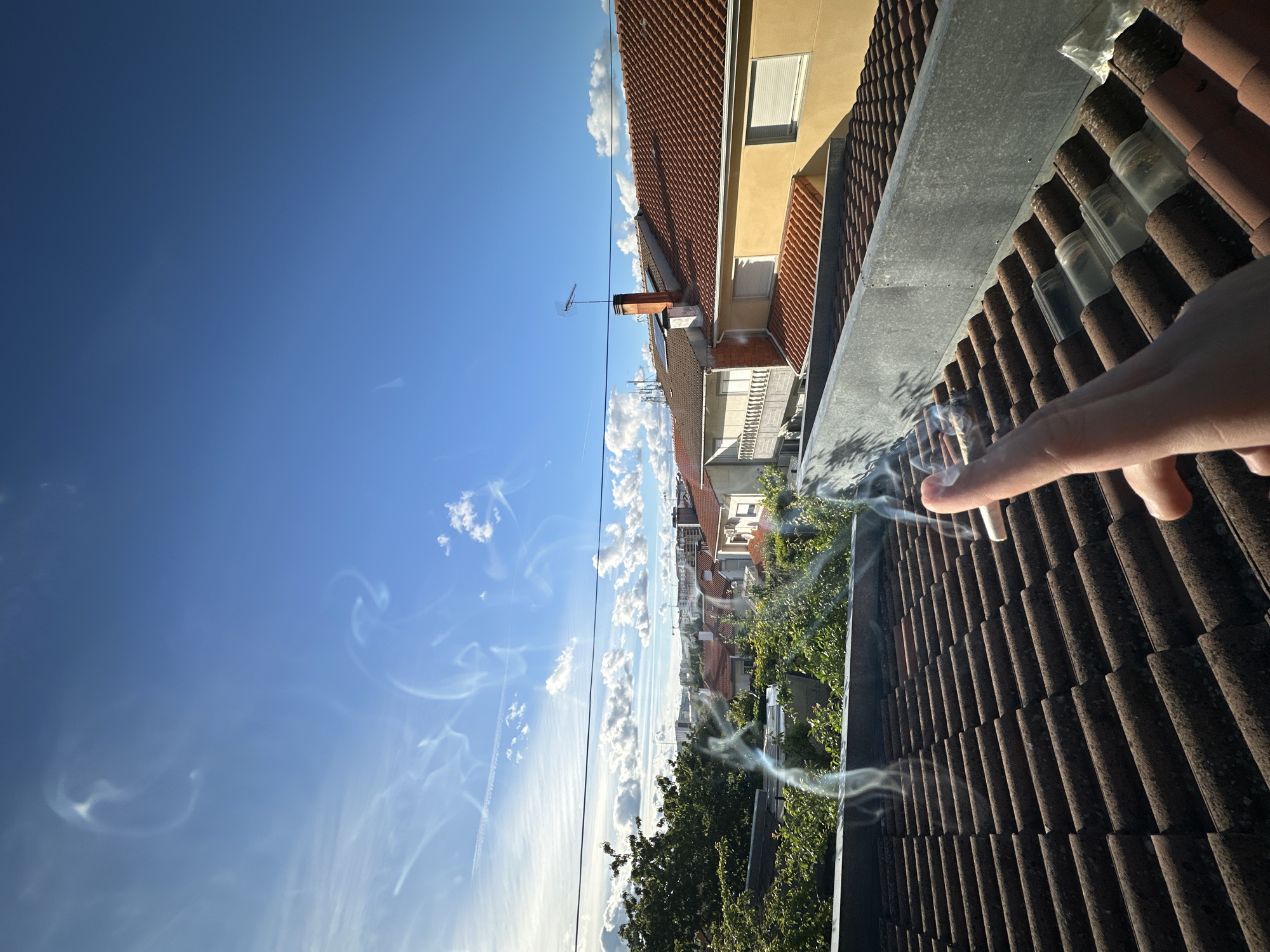 A medium, outdoor shot taken from a high vantage point (possibly a rooftop) features a clear, blue sky with scattered clouds, residential buildings, and a hand holding a cigarette with smoke.
The upper-left quadrant of the photo is dominated by a clear, bright blue sky interspersed with wispy, white clouds. In the upper-right, the roofs of several buildings with red tiles are visible, including the edge of a tan-colored building with a white-slatted window and a balcony. The cityscape in the mid-ground comprises more buildings with red-tiled roofs, interspersed with green foliage, possibly trees in gardens. In the extreme foreground, a pale, human hand holds a white cigarette emitting visible smoke, indicative of burning tobacco. The orientation of the shot is rotated 90 degrees clockwise. Given the architectural style and cityscape, it is possible that the location is in Bragança, Portugal.