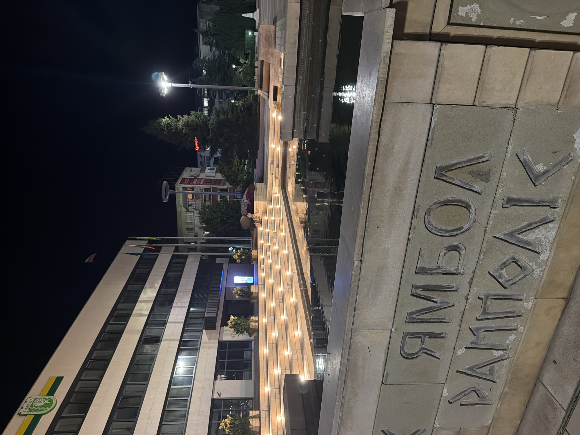 The image depicts a nighttime scene in front of a modern building, likely a municipal building or administrative center, in Yambol, Bulgaria. The building is multi-storied with a facade of windows and is illuminated by artificial lights. In the foreground, there is a water feature with tiered steps, each step illuminated by small, embedded lights, creating a cascading effect. The water reflects the lights and the surrounding architecture. A stone wall in the very foreground features carved text in what appears to be Cyrillic script, with some letters worn. The inscription is divided into two lines, with the upper line possibly reading "ЯМБОЛ" (Yambol) and the lower line partially visible with characters that might spell out another word or name related to the city. Behind the water feature, there are trees, streetlights, and other buildings visible in the distance. The sky is dark, indicating nighttime. The overall impression is one of a well-lit public space at night, possibly a central square or plaza.