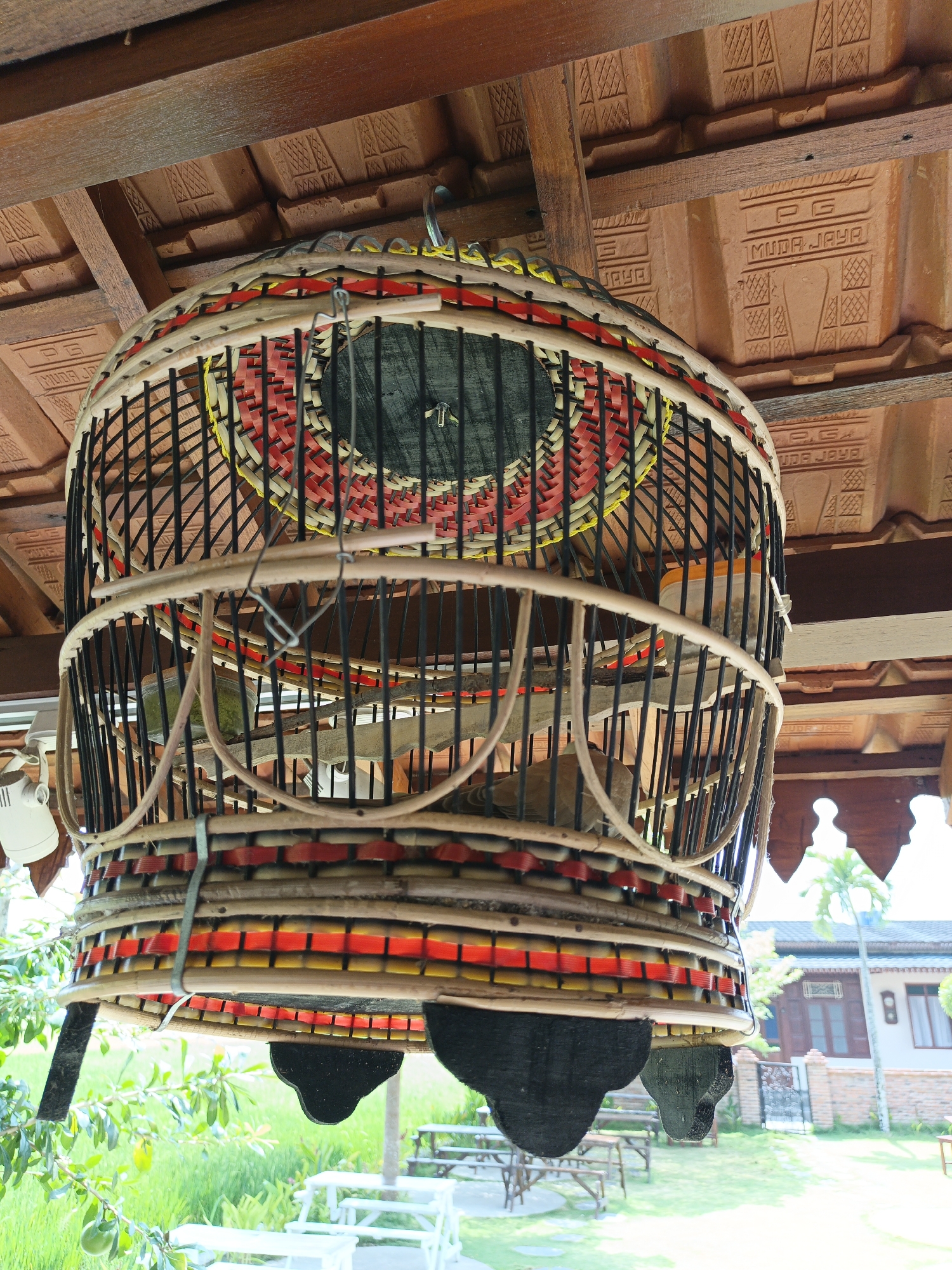 In Sekarsuli, Indonesia, a handcrafted birdcage hangs suspended from a wooden beam beneath a tiled roof. The cage is constructed from woven bamboo and adorned with red and yellow decorative bands. Inside, at least two birds are visible, along with perches and what appears to be a feeding container. The background reveals a tropical outdoor setting with lush green foliage, picnic tables, and a building with a tiled roof and brick accents. The overall scene suggests a tranquil, rural environment.