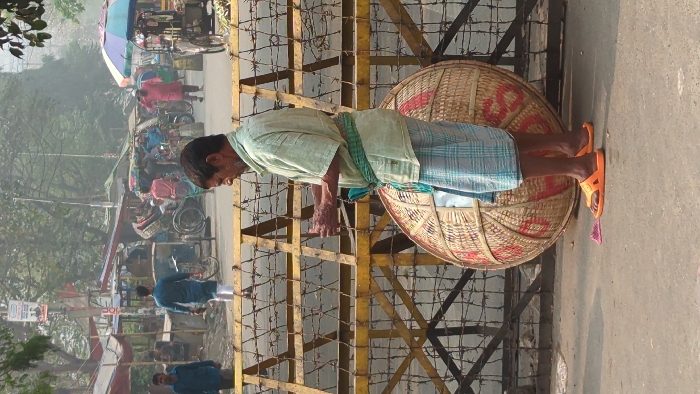 The image depicts an urban street scene in Dhaka, Bangladesh, partially obscured by atmospheric haze. In the immediate foreground, a middle-aged man stands in profile, facing right, wearing a light-colored short-sleeved shirt, a blue and white checkered lungi, a green and white checkered waist sash, and bright orange sandals. He holds his hands up, seemingly manipulating an object. Behind him, resting against a yellow metal barrier, is a large, dome-shaped woven bamboo basket with red markings.
The yellow barrier, composed of metal frames, spans horizontally across the midground and is interwoven with multiple strands of barbed wire, suggesting a blockade or restricted area. Beyond this barrier, a busy street scene unfolds with numerous people and vehicles. Several individuals are visible: a man in a blue shirt and white pants walks away on the left, and another man in a red tunic walks towards the right side of the frame, near a multicolored umbrella. Multiple cycle-rickshaws are present, some parked, others in motion. Makeshift stalls with tarpaulin roofs line the street. The background features sparse trees and utility poles, all under a hazy sky. A small pink object lies on the asphalt ground near the main subject's feet.