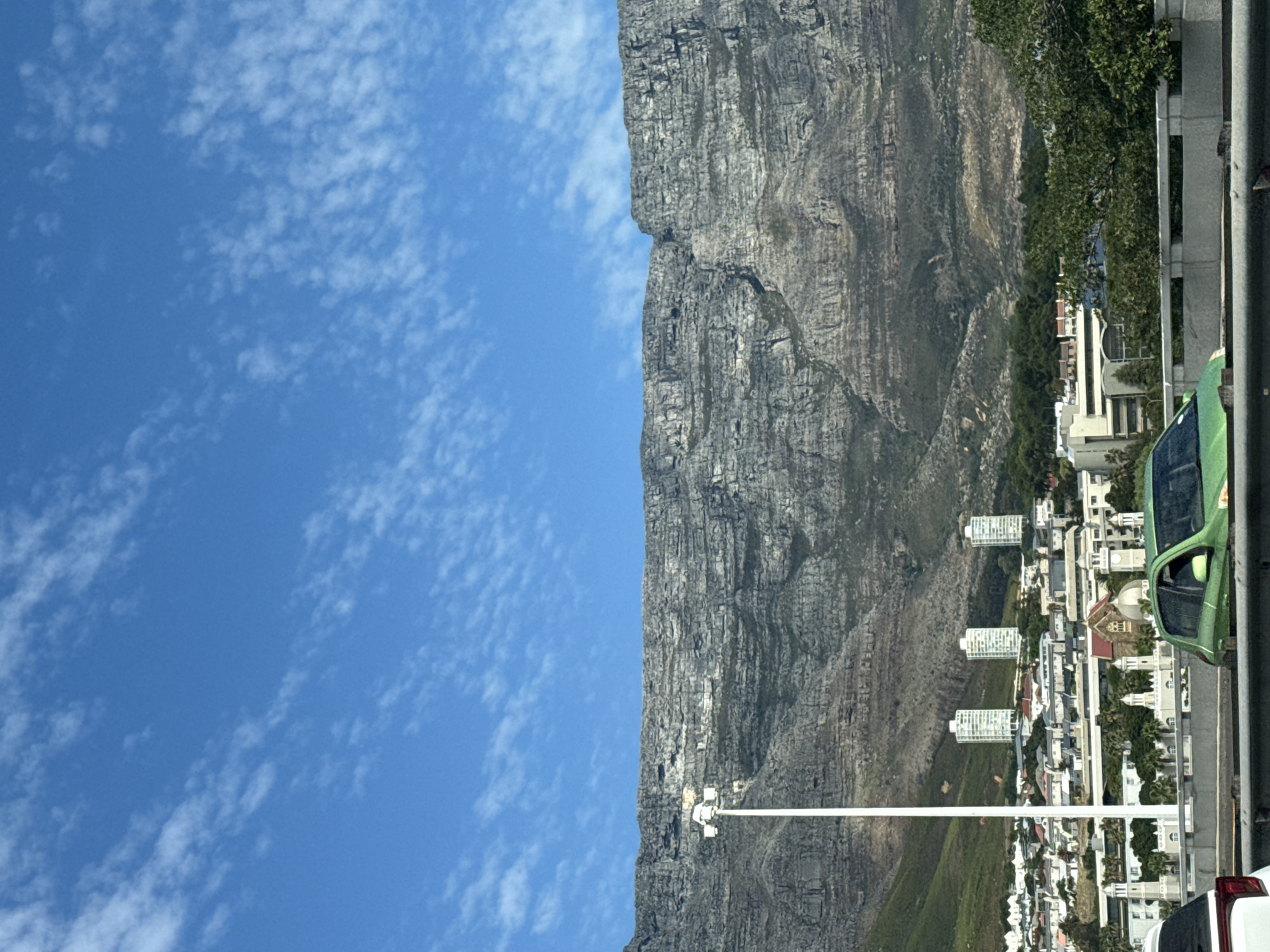 The image shows a view of Table Mountain in Cape Town, South Africa, taken from a street level. To the left is a blue sky with scattered white clouds. Adjacent to the sky is the massive, rocky face of Table Mountain. The mountain's terrain is textured with visible crevices and geological formations.
At the base of the mountain, there is a residential area consisting of houses and buildings. The architecture appears to be modern with some older structures mixed in. A vertical utility pole with a street lamp is also visible. On the right edge of the image, a green car is parked next to a concrete barrier. The rear of another car, white, is also visible.