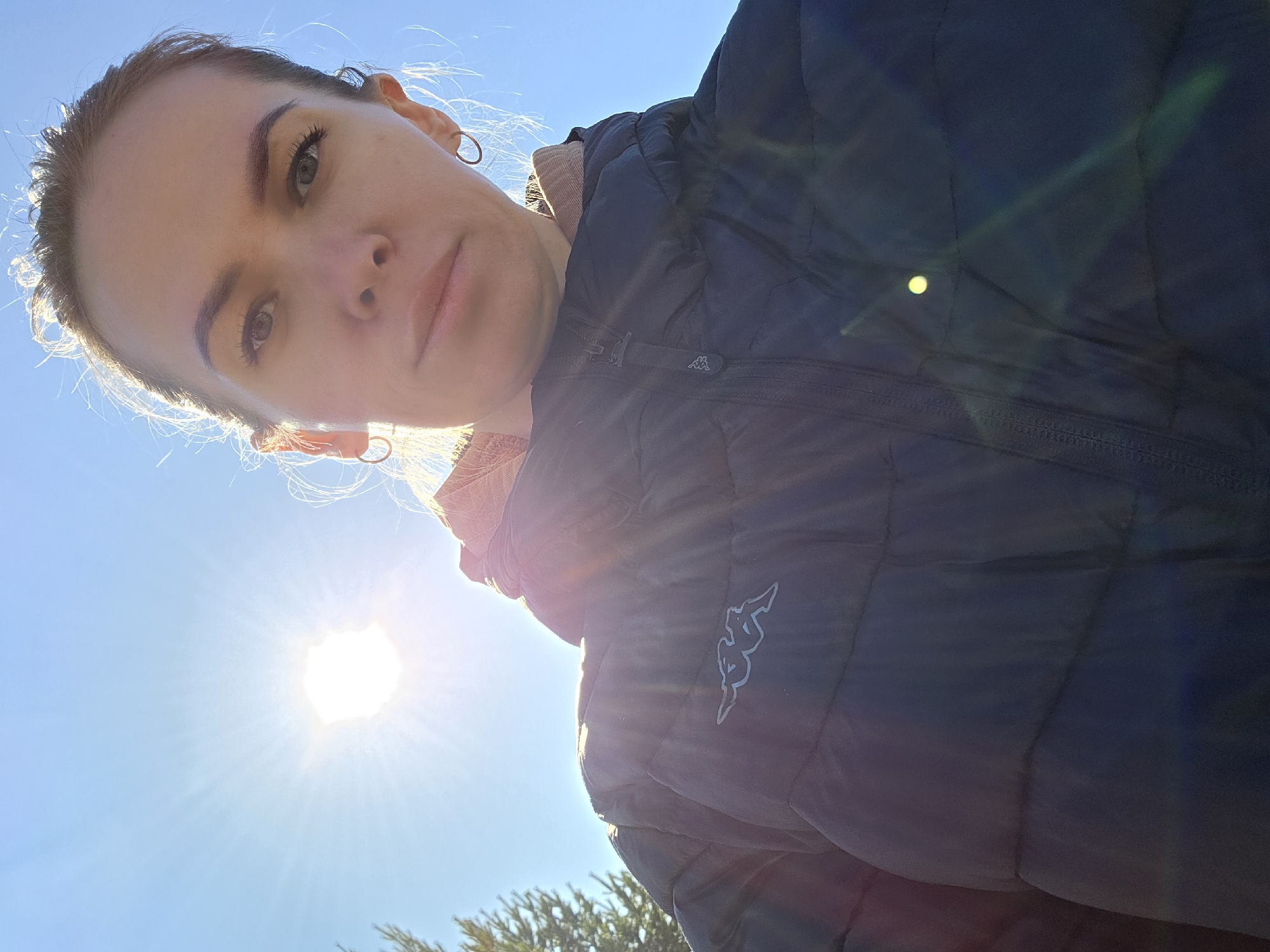 A low-angle selfie of a woman in a puffy blue Kappa jacket. The sun is positioned in the lower left corner of the frame, creating a bright lens flare. The woman has fair skin, light brown hair pulled back, and wears hoop earrings. The environment appears to be outdoors, indicated by a clear blue sky and greenery in the bottom portion of the image. No further details are available to confirm that this picture was taken in Chaikovsky, Russia.