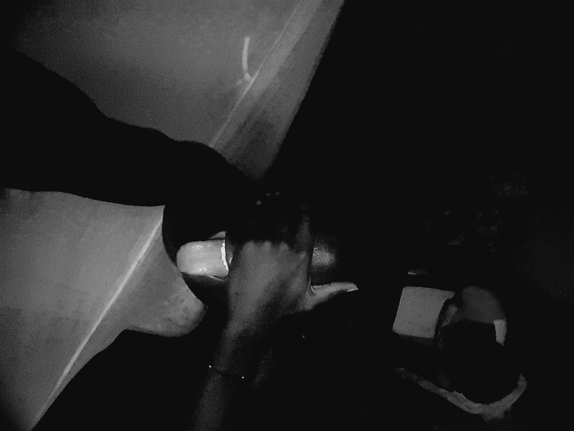 The image is a black and white, low-light, close-up shot of a person's face and hand. The person's eyes are covered by a hand, and their tongue is sticking out. The background is dark and appears to be indoors, with some indistinct shapes that could be furniture or other objects. There is a surface in the foreground that is textured and light in color, possibly a table or a wall. The image is grainy and the lighting is poor, suggesting it was taken in dim conditions, possibly at night. There is no specific indication of the location being Narayi, Nigeria; this detail is external to the visual information provided in the image.