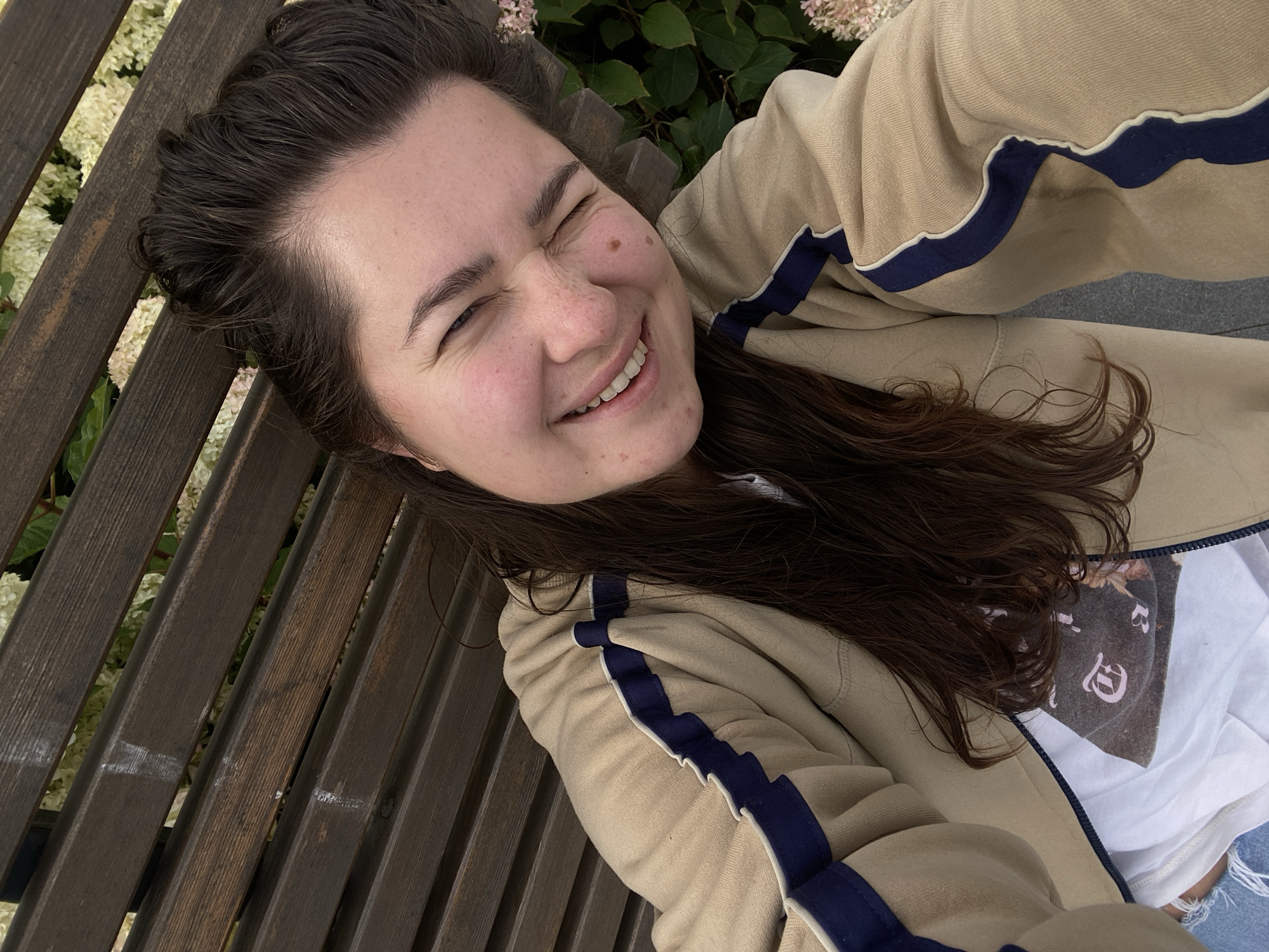A young woman with long, dark brown hair is taking a selfie while sitting on a wooden bench outdoors. She is wearing a beige jacket with blue and white stripes and a white t-shirt with a graphic print. Her eyes are closed, and she is smiling broadly. The background shows lush green foliage with some light-colored flowers and a dark wooden bench. The image does not contain any information about the city of Perm, Russia.