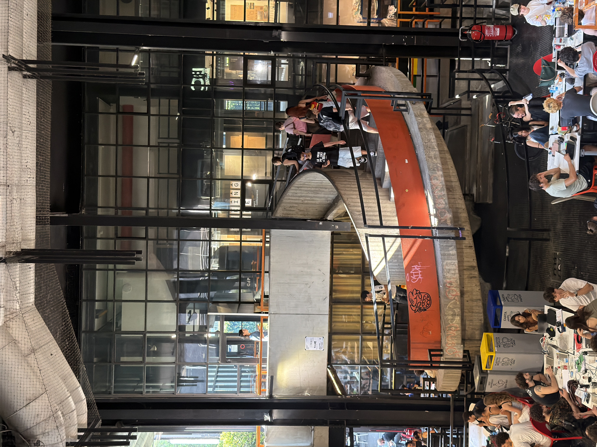 The image depicts a multi-level interior space, likely a public or commercial building in Milan, Italy. The architecture is modern with exposed structural elements, including black metal supports and grid-patterned glass walls. A curved, orange ramp-like structure traverses the scene diagonally, with people walking along it.
On the lower level, multiple individuals are seated at tables, appearing to be working or studying, some using laptops. Recycling bins in yellow and blue are nearby. On the upper levels, individuals are seen walking and congregating. A red fire extinguisher is mounted on a structural support.
The environment is brightly lit and appears to be an active space with a mix of movement and stationary activity. The walls feature graffiti and other markings.