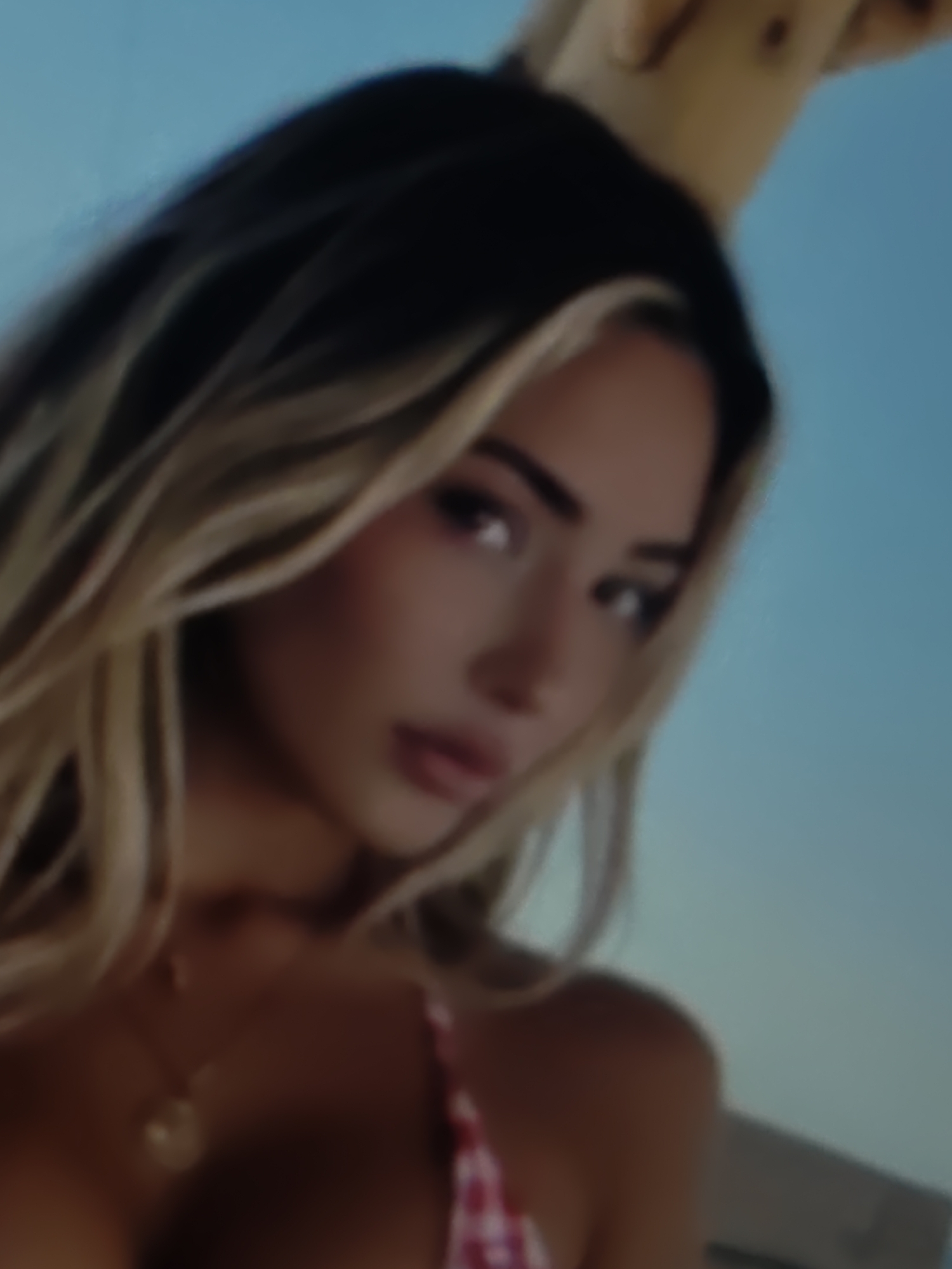 The image is a close-up, blurred shot of a woman with blonde hair and makeup. She is wearing a red and white checkered top and a gold necklace. The background is a clear blue sky. There are no visible elements that would indicate the image was taken in Kurganinsk, Russia.