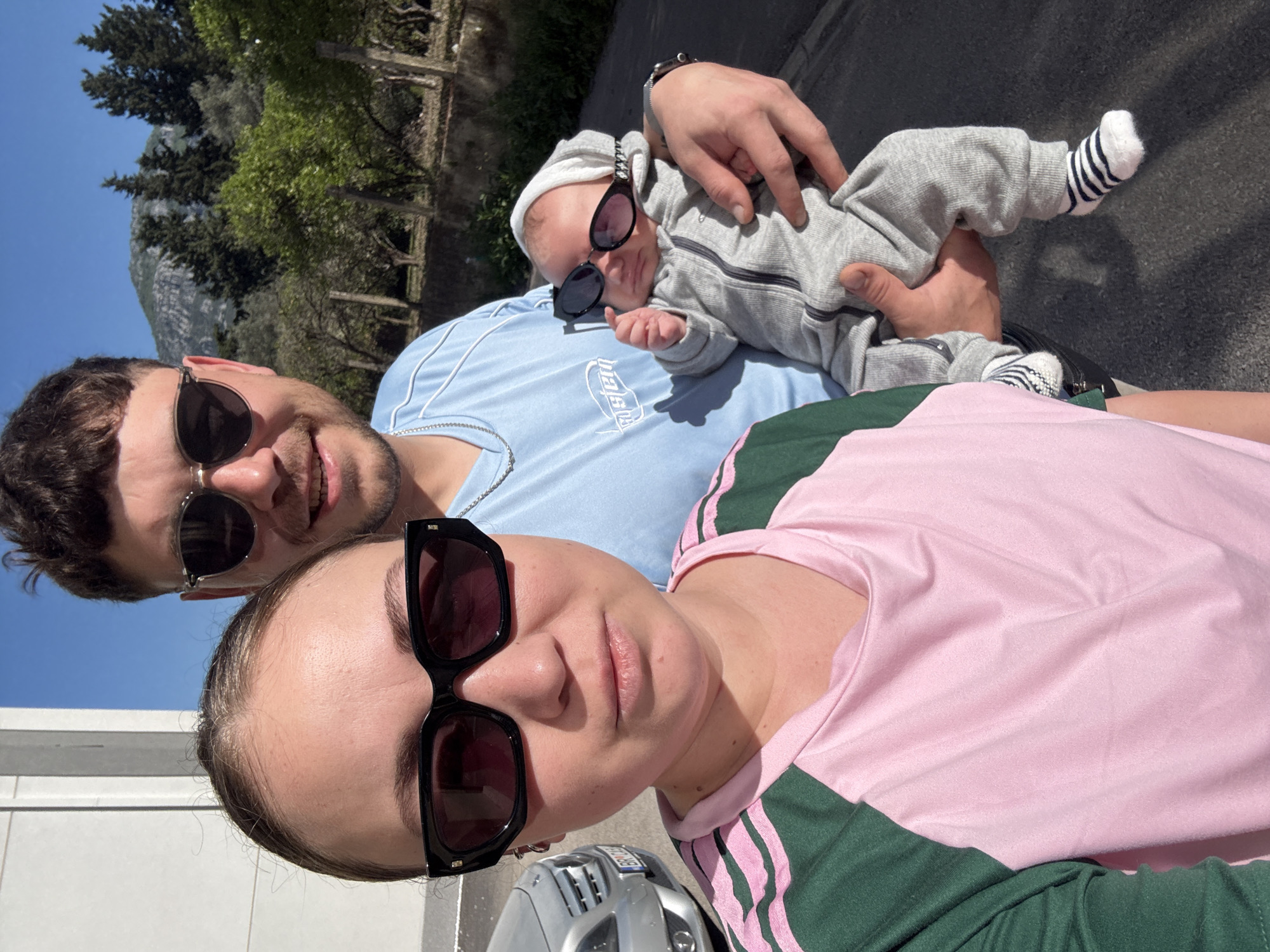 The image depicts a family portrait in Bar, Montenegro, likely taken near a street or sidewalk adjacent to a building and vegetation. The family consists of three individuals: a woman, a man, and an infant. The woman is positioned in the foreground, wearing sunglasses and a pink and green shirt. The man is behind her, wearing sunglasses and a light blue shirt, holding the infant. The infant is also wearing sunglasses and a grey outfit with striped socks. The background reveals a mountainous terrain with trees, suggesting the proximity to the Dinaric Alps. The photograph appears to be taken outdoors in daylight with bright sunshine. The presence of a vehicle is suggested by a partial view of its hood.