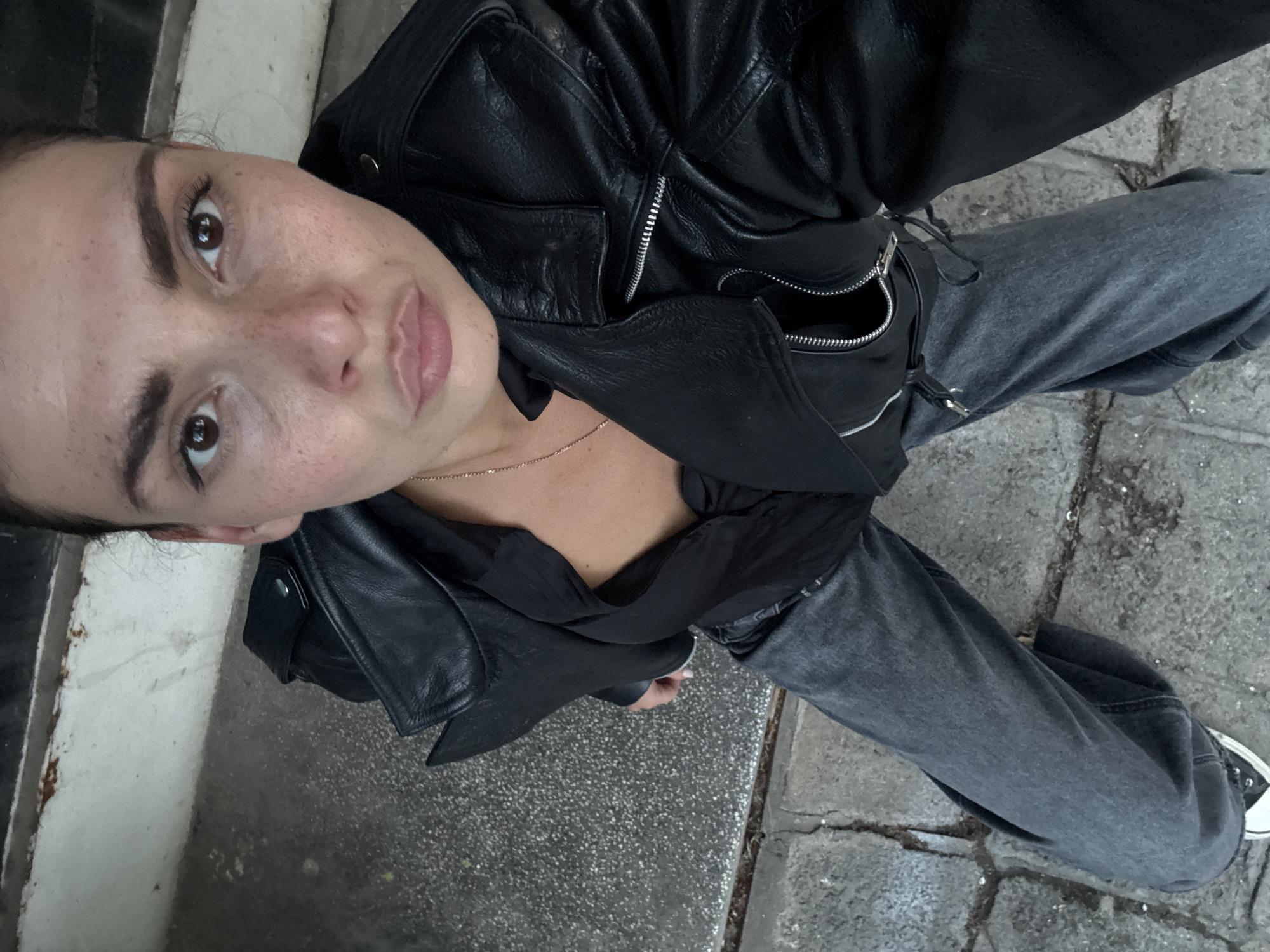 A high-angle, overhead selfie captures a person standing outdoors in Sofia, Bulgaria. The subject, who appears to be female, looks directly up at the camera with brown eyes and a neutral expression. Her face is lightly freckled, with defined eyebrows and natural-toned lips.
She is dressed in a stylish black leather jacket featuring silver zippers and hardware, worn over a dark, possibly black, top that reveals a delicate gold-colored chain necklace. Her lower body is clad in dark grey jeans or trousers, which appear relaxed fit. On her right foot, a black and white sneaker is visible.
The setting is an urban outdoor environment, with the ground paved in a mix of concrete and possibly cobblestones, showing signs of wear and cracks. A distinct white curb or painted line is visible along the left edge of the frame. The lighting indicates it is daytime. The overall impression is a casual, contemporary street style portrait. No text is visible in the image.
