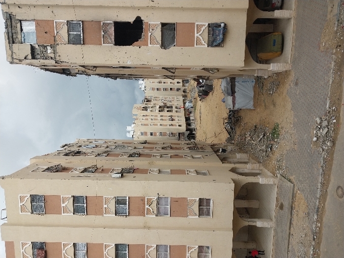 This image depicts a scene in the city of Khan Yunis, Palestinian Territory, characterized by residential buildings showing significant damage and debris-strewn ground.
Two multi-story, light-tan apartment buildings with brown window trim frame a narrow, sandy open area filled with rubble. The building on the left exhibits numerous small pockmarks across its facade and some minor window damage, but is largely structurally intact. Its ground floor features a series of arched openings, underneath which two individuals, one in a red shirt appearing to be a child, are partially visible. The building on the right displays severe damage, particularly on its upper floors, where large sections of concrete are missing, exposing dark interiors and twisted rebar. Its facade also shows multiple small pockmarks. The ground floor of this building also has arched openings, with a green tarp-covered object, possibly a vehicle, parked in one bay, and part of another vehicle visible adjacent to it.
The central ground between these two buildings is unpaved, consisting of sand and dirt heavily intermingled with concrete rubble, twisted metal, and discarded materials. A makeshift shelter, constructed from white tarpaulin and other coverings, stands on the right side of this central area. In the background, more multi-story residential buildings of similar architecture are visible, some also appearing damaged, under an overcast sky. A faint, broken cable stretches horizontally between the two prominent foreground buildings. The immediate foreground features a grey paved area of interlocking pavers, with small piles of debris near its edge and a manhole cover visible on its surface.