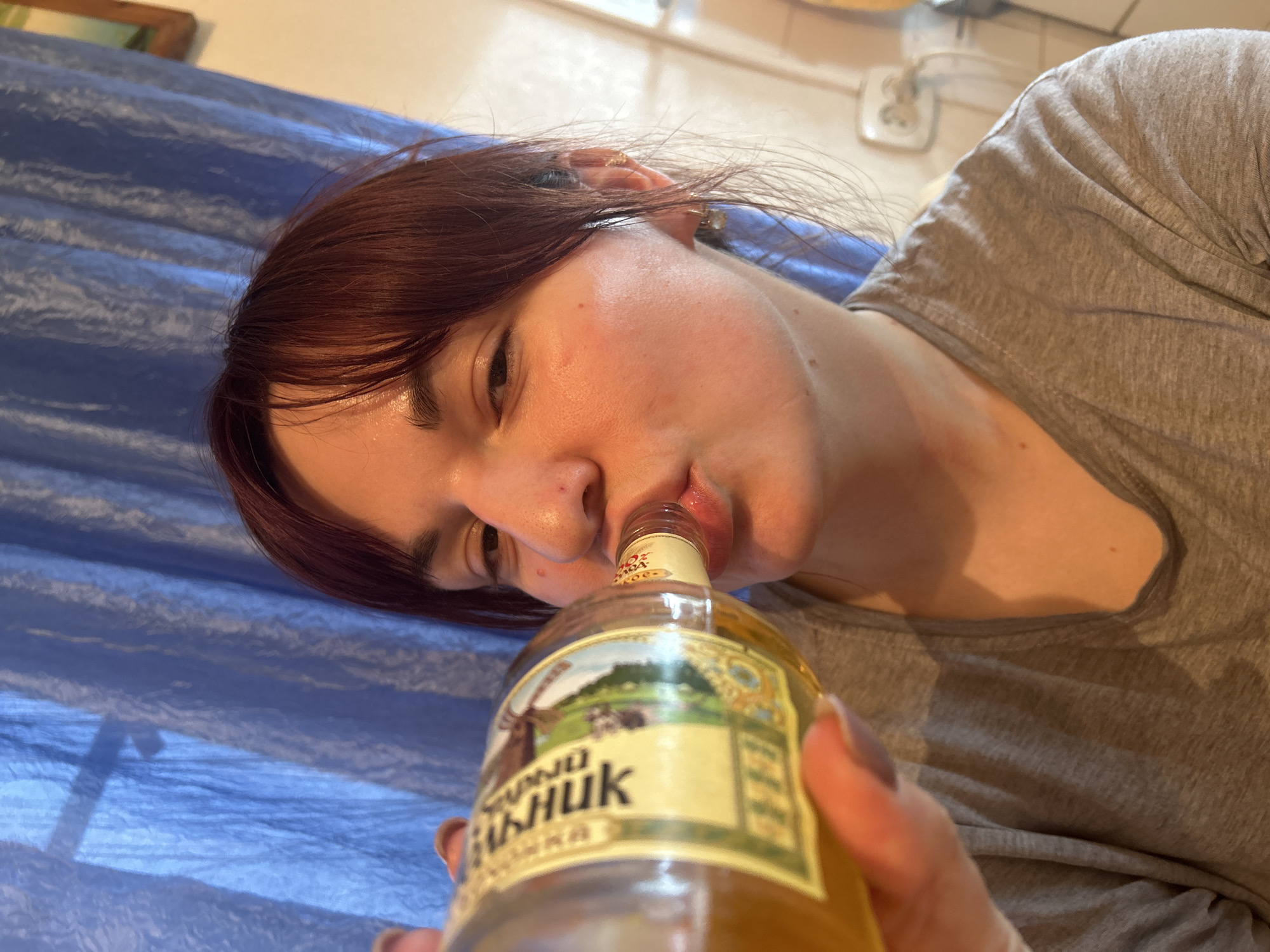 A low-angle, close-up shot captures a person, likely a young woman with reddish-brown hair, drinking directly from a glass bottle. Her face is turned towards the camera, with her mouth on the bottle's opening. Her eyes are partially obscured or downcast. She is wearing a grey t-shirt and appears to be lying down on a surface covered by a rumpled blue sheet or blanket. The lighting creates a slight sheen on her skin, possibly indicating sweat or oil.
The glass bottle contains a light amber-colored liquid. Its label, featuring a landscape graphic with a windmill, displays prominent Cyrillic text. Visible words include "БАШКИРСКИЙ СТАНДАРТ" (Bashkir Standard) and "КОНЬЯК" (Cognac). Other visible text reads "СТАРЫЙ" (Old) and "СЫНЖИК".
The background shows a light-colored wall with an electrical outlet or switch, suggesting an indoor setting, possibly a casual home environment. The location context for this image is Foki, Russia. The image captures a candid moment of consumption.