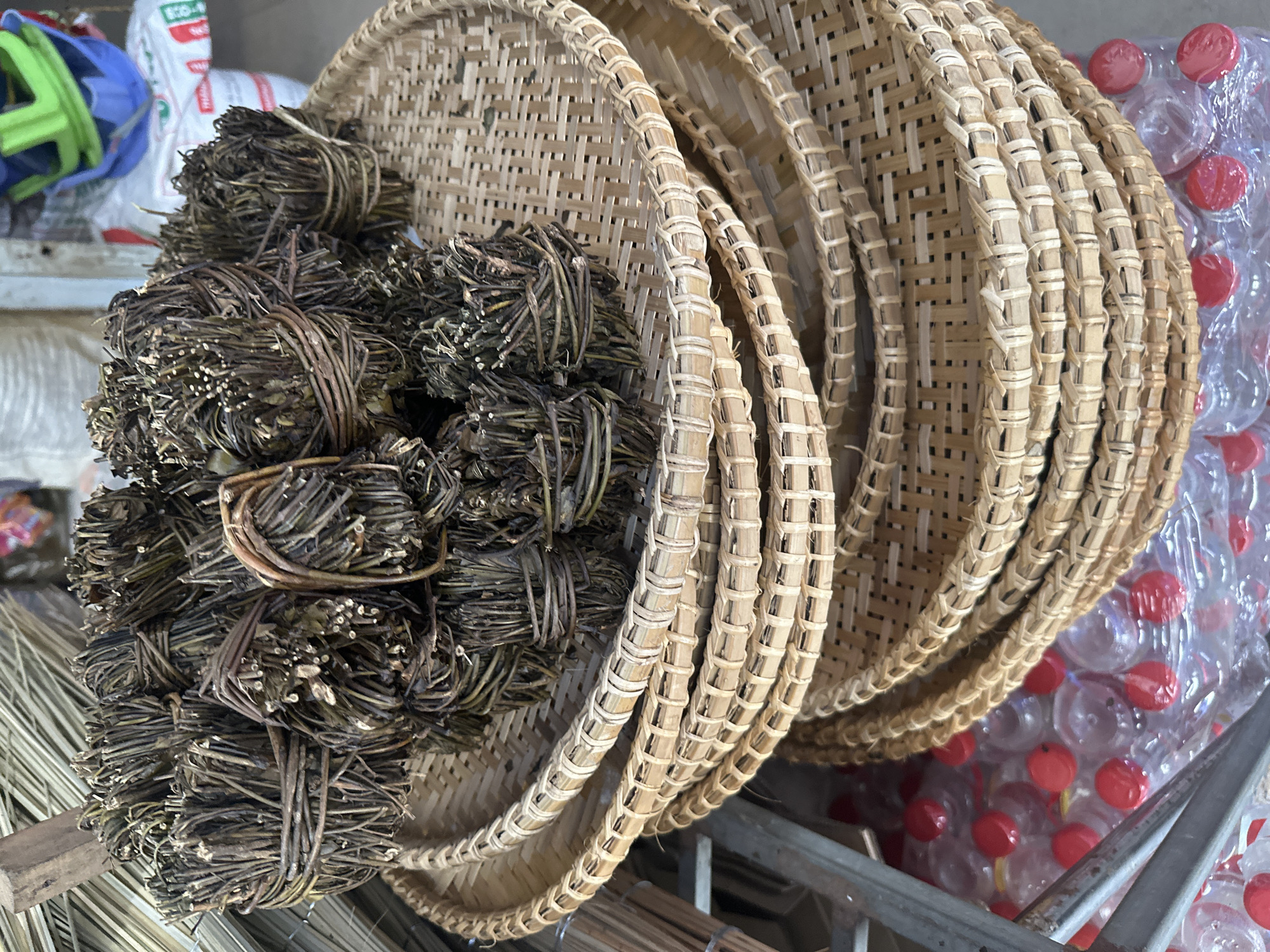 The image shows a stack of woven bamboo trays next to bundles of dried herbs or plant material. The trays are round, with a light-colored woven base and a thicker, braided rim. Several bundles of dried herbs are stacked on top of one of the trays. To the right of the trays, there is a package of clear plastic bottles with red caps. The setting appears to be a market or storage area in Bac Nha Trang Ward, Vietnam.
