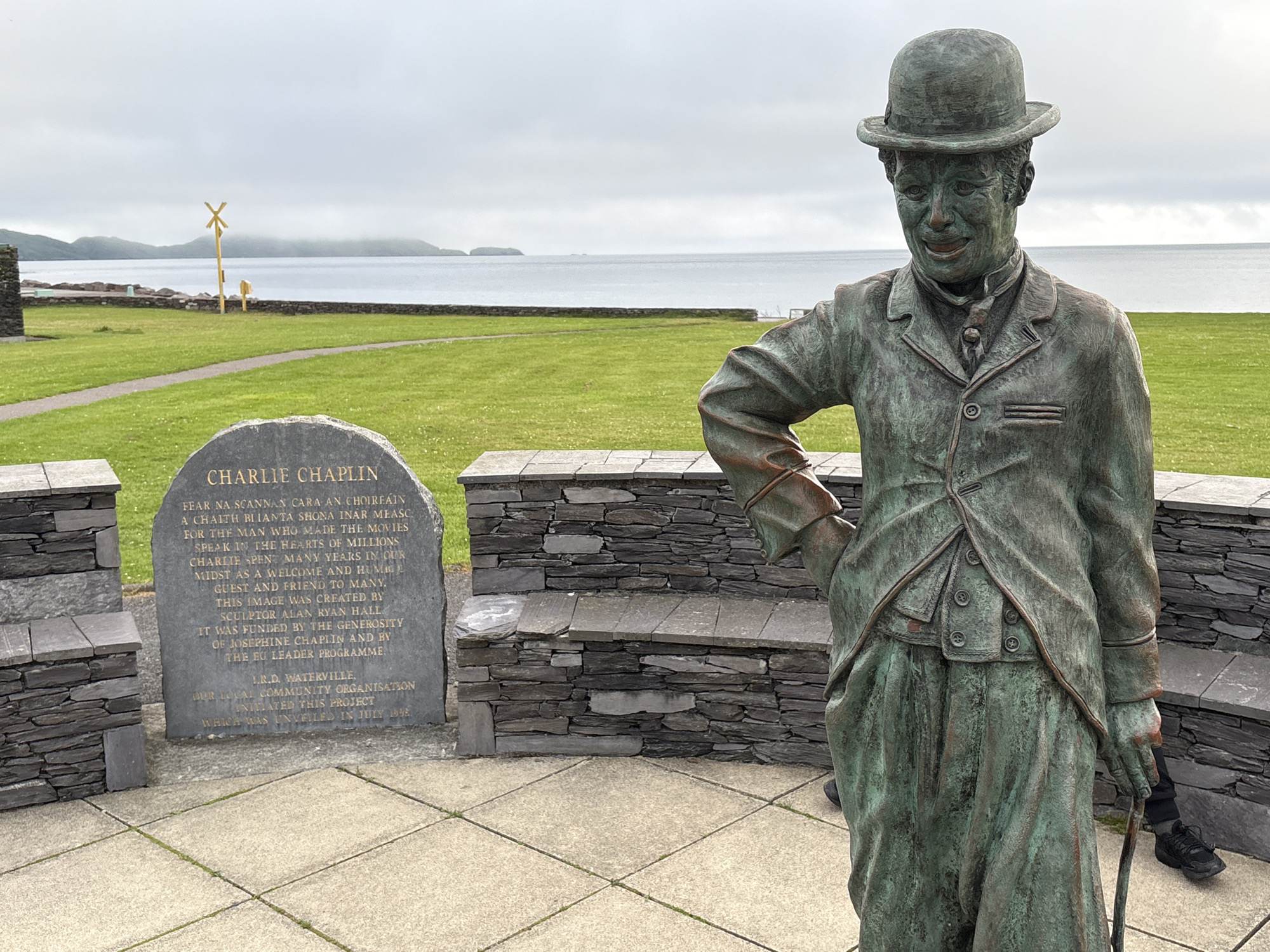 This media file features a bronze statue of Charlie Chaplin in Waterville, Ireland. The statue depicts Chaplin in his iconic Tramp persona, complete with a bowler hat, suit, and cane. He is positioned in the foreground, looking slightly to the left, with a stone monument to his left and a stone wall with a bench behind him.
The monument is inscribed with text honoring Charlie Chaplin, detailing his connection to the area and acknowledging the creators and funders of the statue. Visible text includes "CHARLIE CHAPLIN," "FEAR NA SCANNAN CARA AN CHOIREAIN A CHAITH BLIANTA SHONA INAR MEASC FOR THE MAN WHO MADE THE MOVIES SPEAK IN THE HEARTS OF MILLIONS CHARLIE SPENT MANY YEARS IN OUR MIDST AS A WELCOME AND HUMBLE GUEST AND FRIEND TO MANY. THIS IMAGE WAS CREATED BY SCULPTOR ALAN RYAN HALL IT WAS FUNDED BY THE GENEROSITY OF JOSEPHINE CHAPLIN AND BY THE EU LEADER PROGRAMME. I.R.D. WATERVILLE, OUR LOCAL COMMUNITY ORGANISATION INITIATED THIS PROJECT WHICH WAS UNVEILED IN JULY 1998."
The setting is outdoors, with a grassy lawn stretching towards a body of water, likely the Atlantic Ocean, and hills in the distance. The sky is overcast, suggesting a cloudy or misty day. The overall mood is one of remembrance and tribute. The statue is a prominent object in the scene, serving as a focal point for visitors. The presence of the monument and the statue together creates a memorial space dedicated to the beloved entertainer.