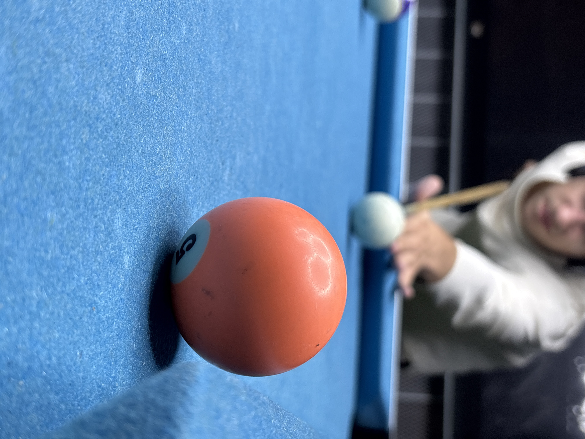 A close-up shot features a pool table with a solid orange ball resting against the rail. The ball has the number "5" visible. An individual, partially out of focus, is holding a pool cue, seemingly about to strike a white ball positioned on the table. The table is covered in blue felt. The background suggests an indoor recreational area. The city of Baan Bang Rak, Thailand is not directly discernible from the image.