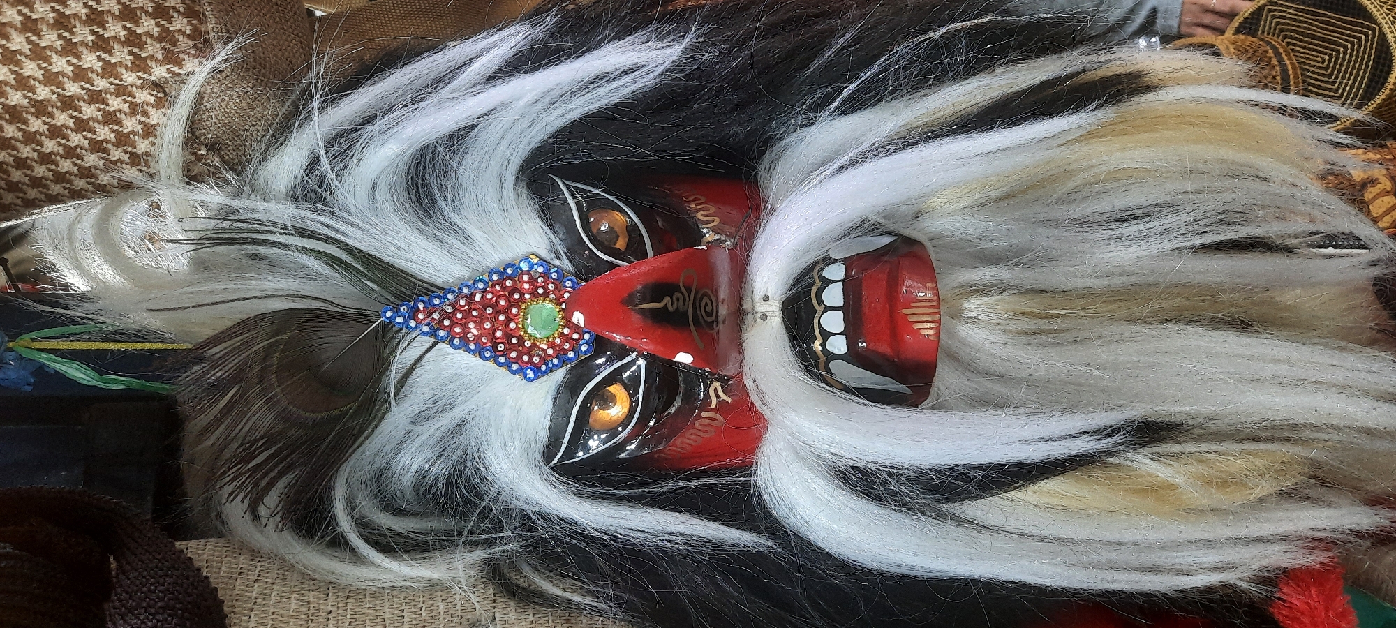 The image is a close-up, head-on shot of a mask with a fierce expression. The mask has large, orange eyes with black pupils and black outlines. The face is primarily red, with black markings around the eyes and nose. The nose is a prominent red structure with swirling gold markings on it. The mouth is open, revealing white teeth and fangs, with the tongue also painted red. A large white beard and prominent white eyebrows frame the face. Black and white hair, made of what appears to be synthetic fibers or animal hair, sprouts from the top and sides of the head, with a few peacock feathers visible near the top. A decorative headdress with red and blue sequins and a green jewel is centered on the forehead. The background is blurred, but appears to be composed of textured fabrics in brown and beige tones, suggesting the mask may be displayed or stored. There are no visible people or specific environmental details that would indicate a location within the city of Blitar, Indonesia.