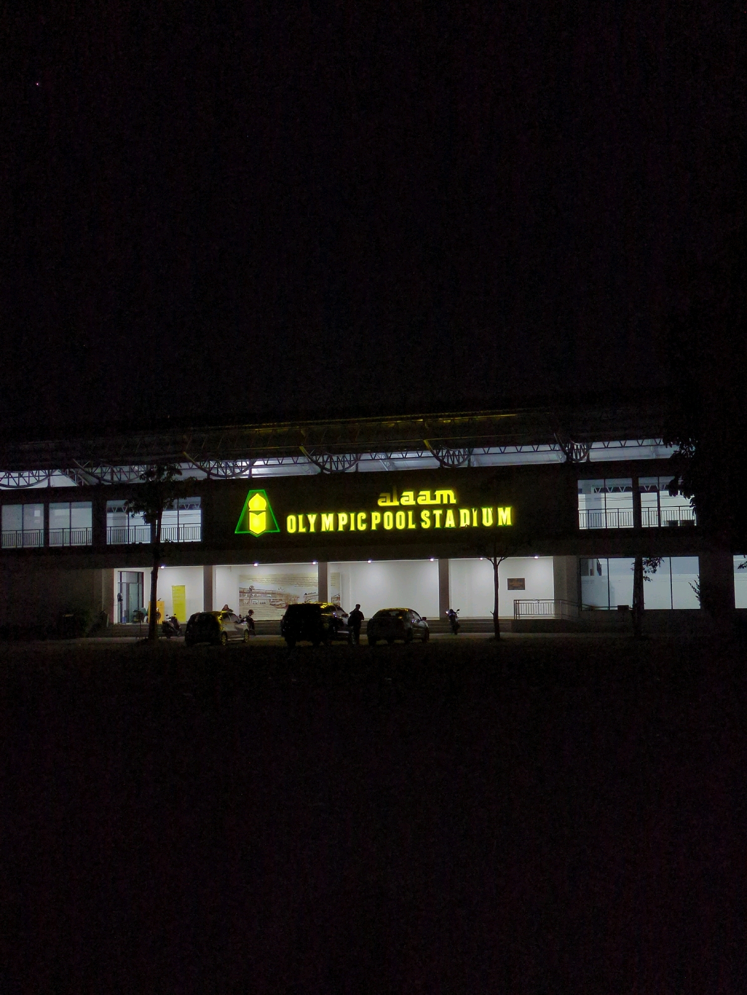 The image depicts the "a.a.m. OLYMPIC POOL STADIUM" at night. The stadium's facade is illuminated by neon yellow signage. To the left of the sign, there is a green triangular logo. The building has multiple levels with windows and balconies. In front of the stadium, several cars and motorcycles are parked. A few people are visible near the vehicles and entering or exiting the building. The environment is dark, suggesting it is nighttime, with the stadium being the primary source of light. The scene is located in Pabelan, Indonesia.