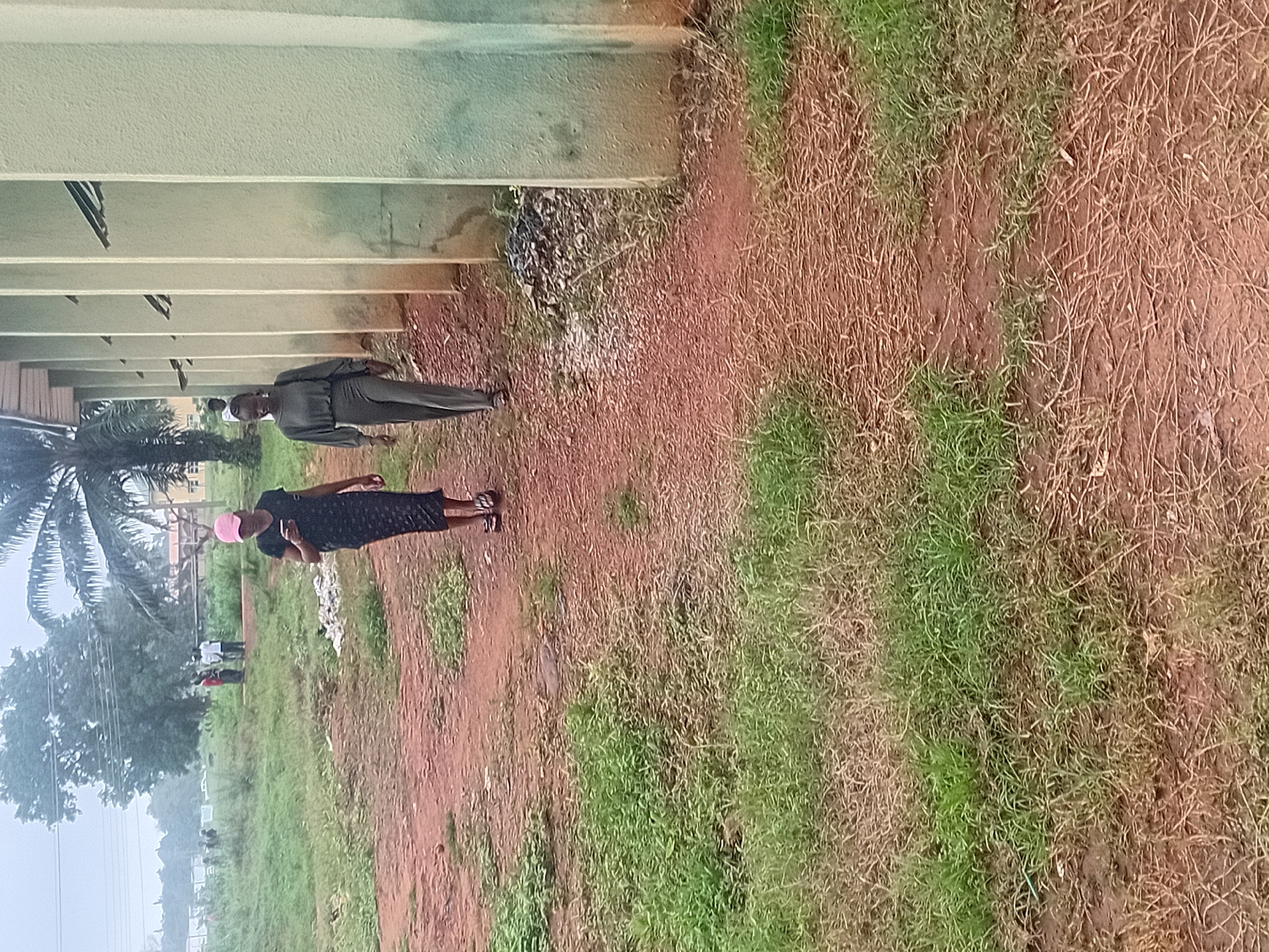 In Ogbogbo, Nigeria, two young women are walking on a dirt path beside a building with light green pillars. The woman on the left wears a dark patterned dress and a pink baseball cap, and she is looking at her phone. The woman on the right is dressed in a gray two-piece outfit and carries a dark bag over her shoulder. In the background, several people are visible standing near trees and other structures. The ground is covered with reddish-brown dirt and patches of green grass. The sky is overcast, suggesting a cloudy or rainy day.
