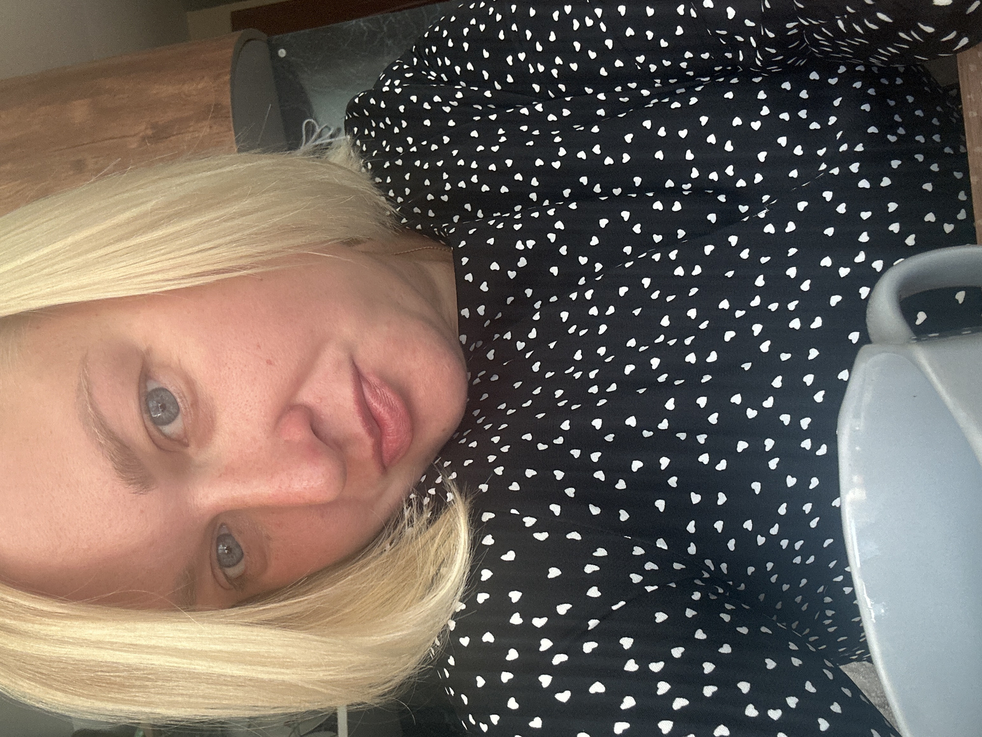 A medium close-up, head and shoulders shot shows a fair-skinned woman with blonde hair and light blue eyes posing for a selfie. She is wearing a black shirt with a white heart pattern and is holding a gray mug. The background appears to be a kitchen, with wooden cabinets and a countertop visible. The scene is indoors and lit by natural light. The woman's expression is neutral, with her lips slightly parted. There are no other people or animals visible in the image. This image does not provide enough information to determine its location in the city of Chaikovsky, Russia.