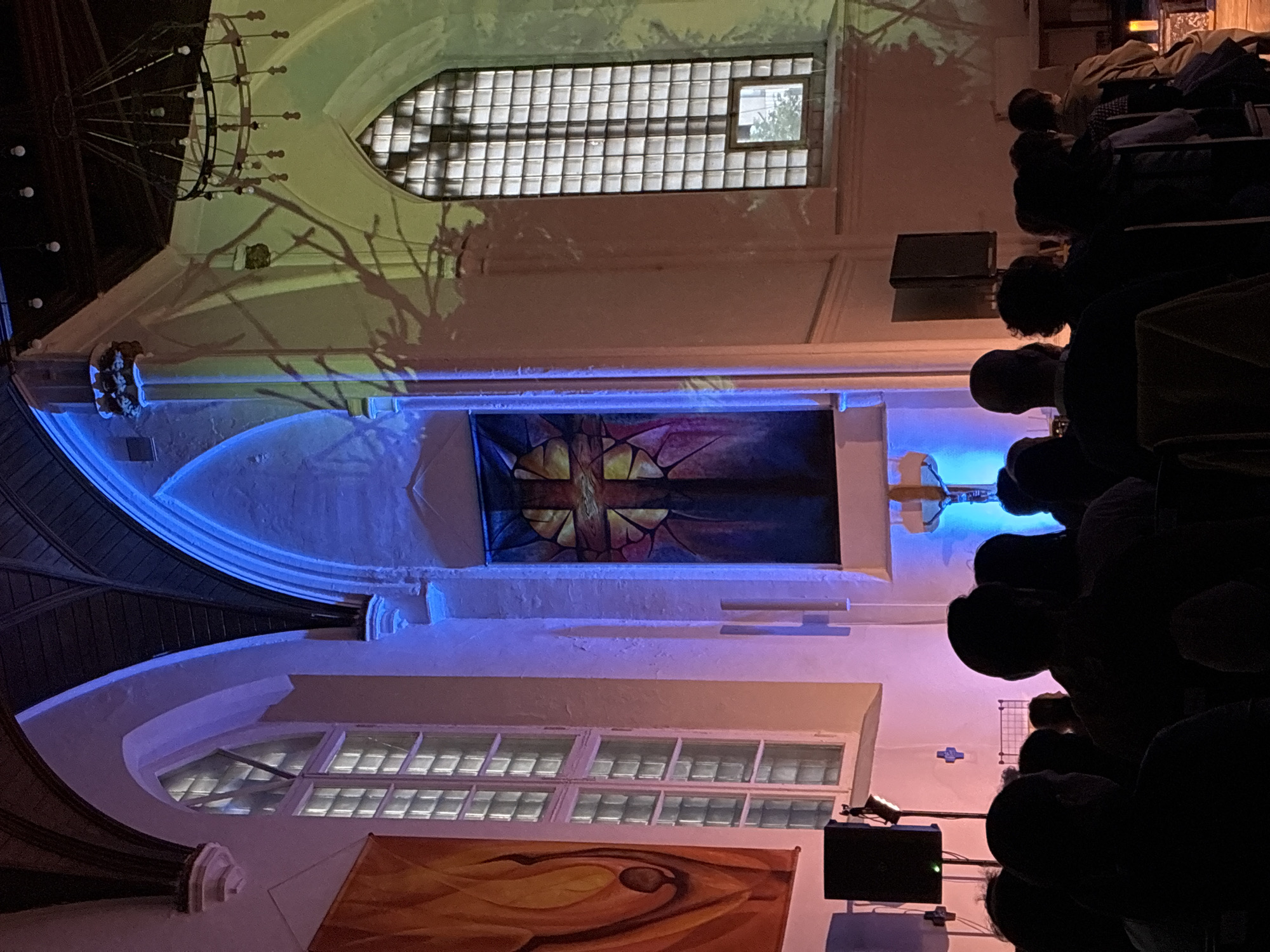 The image shows an indoor scene with a gathering of people, likely an audience, seated in rows on the right side of the frame. The interior appears to be that of a church or similar religious venue, indicated by the architecture including arched ceilings, stained glass windows, and decorative banners/tapestries on the walls. A large chandelier is visible at the top left. The lighting is dramatic, with blue and purple hues dominating the scene. There is a speaker on the lower right, and some indistinct objects at the top. The location is potentially in Moscow, Russia based on architectural styles and possible cultural cues.