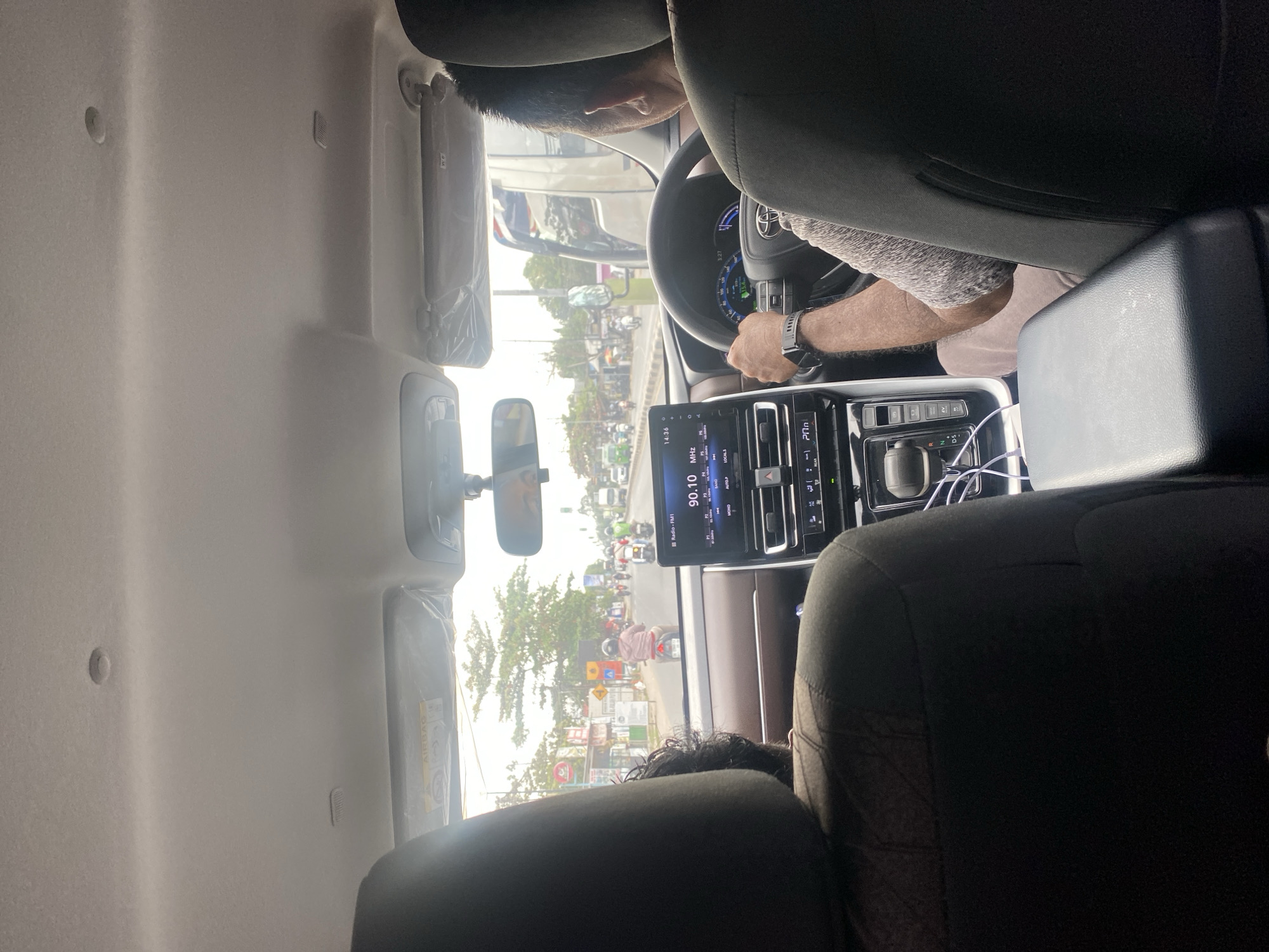 The image depicts the interior of a car driving on a road in Banjarbaru, Indonesia. The view is from the back seat, looking forward. The driver, a man with short hair and wearing a grey and white patterned shirt, is steering the vehicle. His left hand is on the steering wheel, and his right arm is resting. A black watch is visible on his left wrist. The car's dashboard features a digital display showing the radio station "90.10 MHz" and the time "14:36." Gear selector, buttons, and a white USB cable are also visible on the center console.
A second passenger, partially visible from the back, is seated in the front passenger seat. The car's interior is detailed with brown and black upholstery. Outside the car, the scene is a busy street with other vehicles, including motorcycles, and a truck. Pedestrians are also present. Buildings and trees line the roadside, suggesting an urban or suburban environment. The sky is overcast.