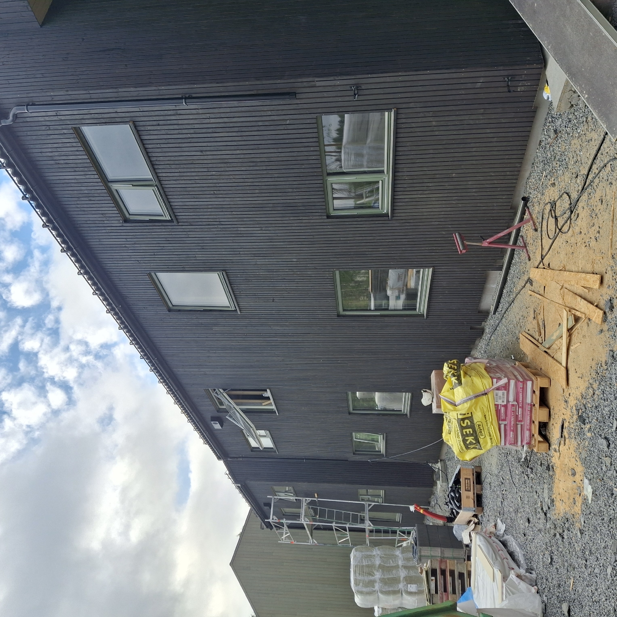 The image depicts a construction site in Teigebyen, Norway. A dark-clad, multi-story building with vertical wooden planks is the primary structure. Several windows are visible, some open. A metal scaffolding system is erected next to the building, with ladders and platforms. In the foreground, construction materials are stacked on pallets, including bags of insulation and other building supplies. Various tools and debris are scattered across the gravel-covered ground. A large yellow bag with "ISEKI" branding is prominently displayed. The sky is cloudy with patches of blue.