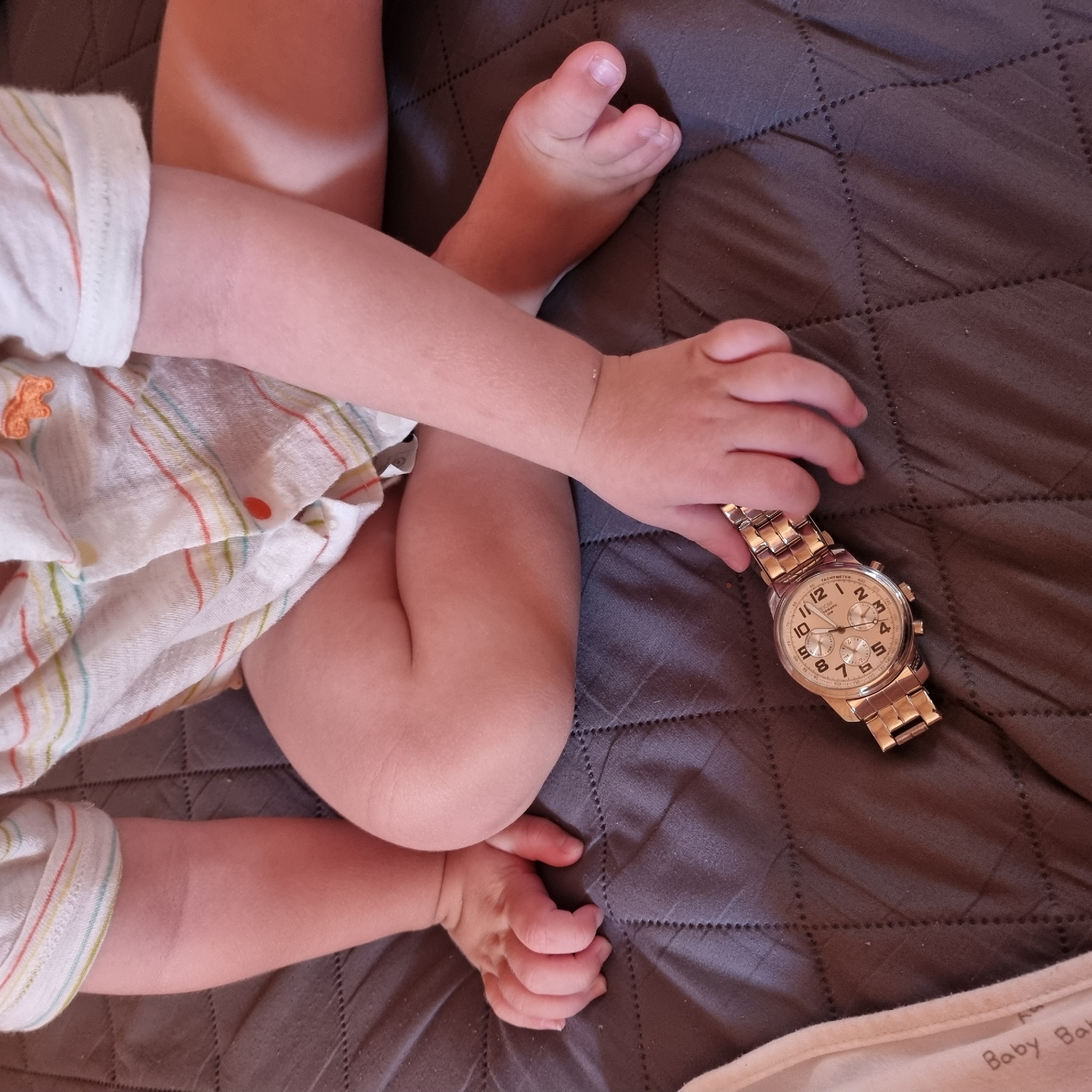 An infant, wearing a striped shirt, sits on a dark gray quilted surface. The infant's hands are resting on the surface, and a gold-colored wristwatch is positioned in front of the infant's right hand. The infant's legs are bent and positioned in front of its body. The background is indistinct. This image was taken in Dunnae-myeon, South Korea.