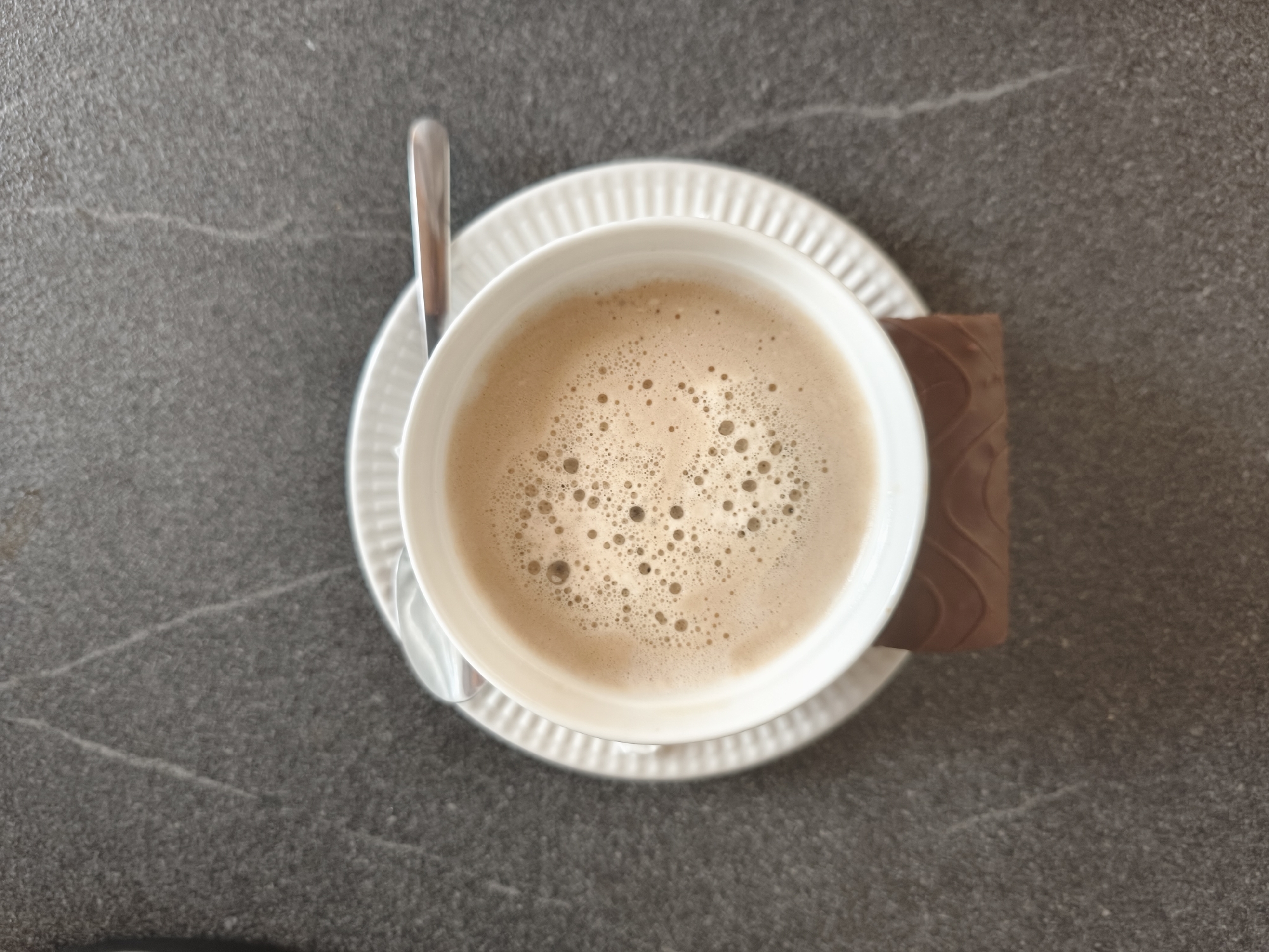 A top-down view shows a white ceramic cup filled with frothy, light brown coffee on a white fluted saucer. A silver spoon rests on the saucer to the left of the cup. To the right of the cup, a rectangular piece of dark chocolate with a patterned surface sits on the saucer. The scene is set against a dark gray speckled countertop, possibly granite or a similar material, with subtle lighter gray veining. The lighting suggests an indoor setting, possibly a cafe or a home. There are no people visible in the image.