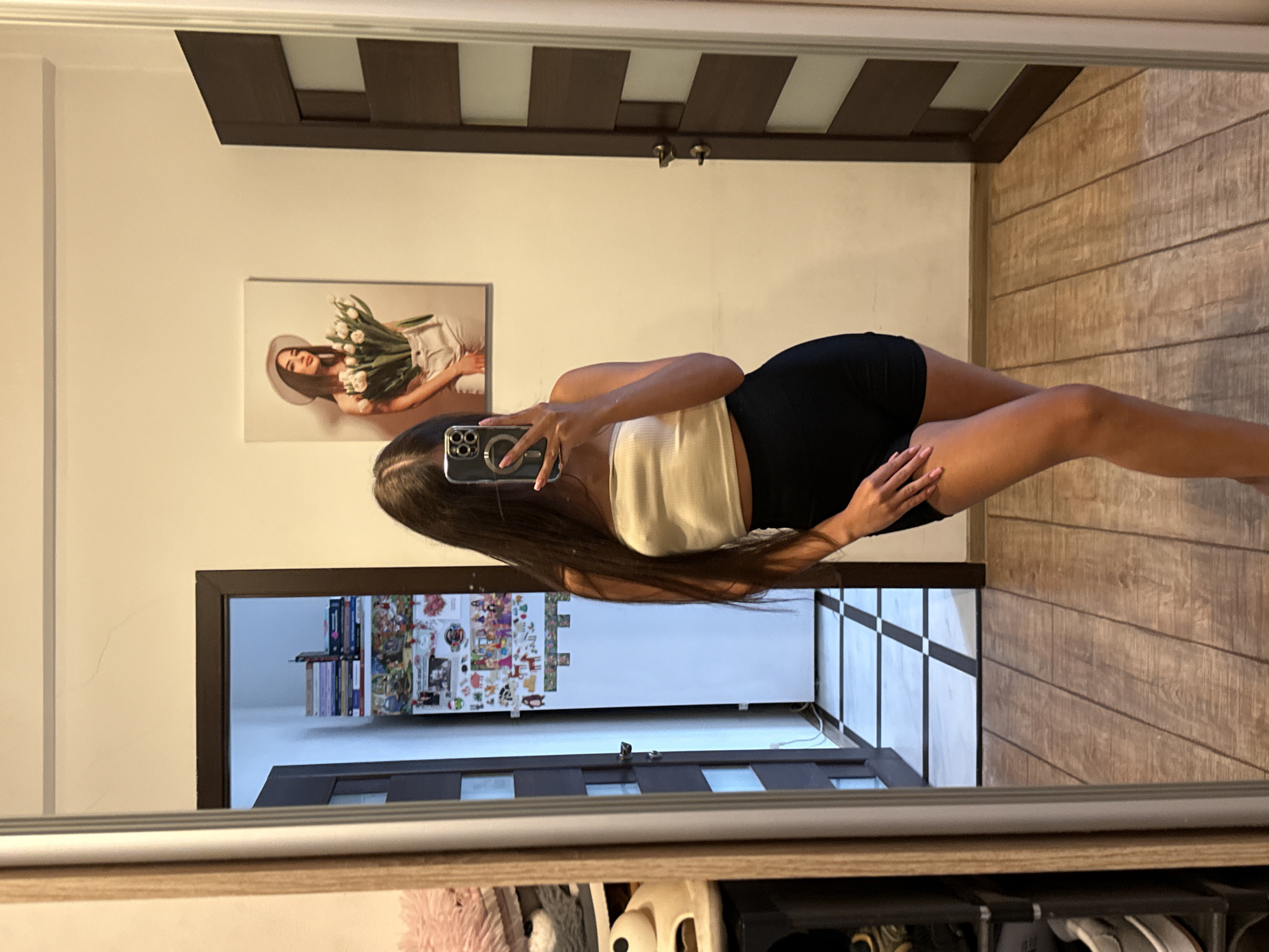 A young woman with long dark hair is standing in front of a full-length mirror, taking a selfie. She is wearing a white strapless top and black shorts. The mirror is in a room with wooden floors and cream-colored walls. To the left of the mirror, there is a closet with shelves containing various items, including shoes and a pink fluffy object. In the background, a door opens to a hallway with tiled floors. On the wall opposite the mirror, there is a framed picture of a woman holding a bouquet of white tulips. The scene appears to be in a residential setting, likely an apartment or house, within Chișinău, Moldova.