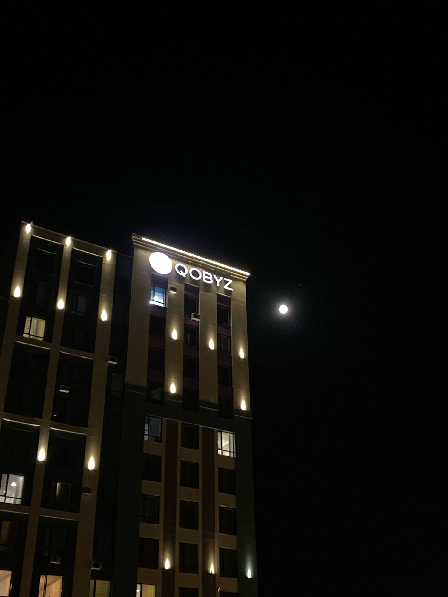 A night view of the Qobyz hotel building in Astana, Kazakhstan, features its illuminated signage and architectural lighting. The building is tall and modern, with multiple floors and windows. A full moon is visible in the dark sky to the right of the building. The overall scene is serene and evokes a sense of urban tranquility.