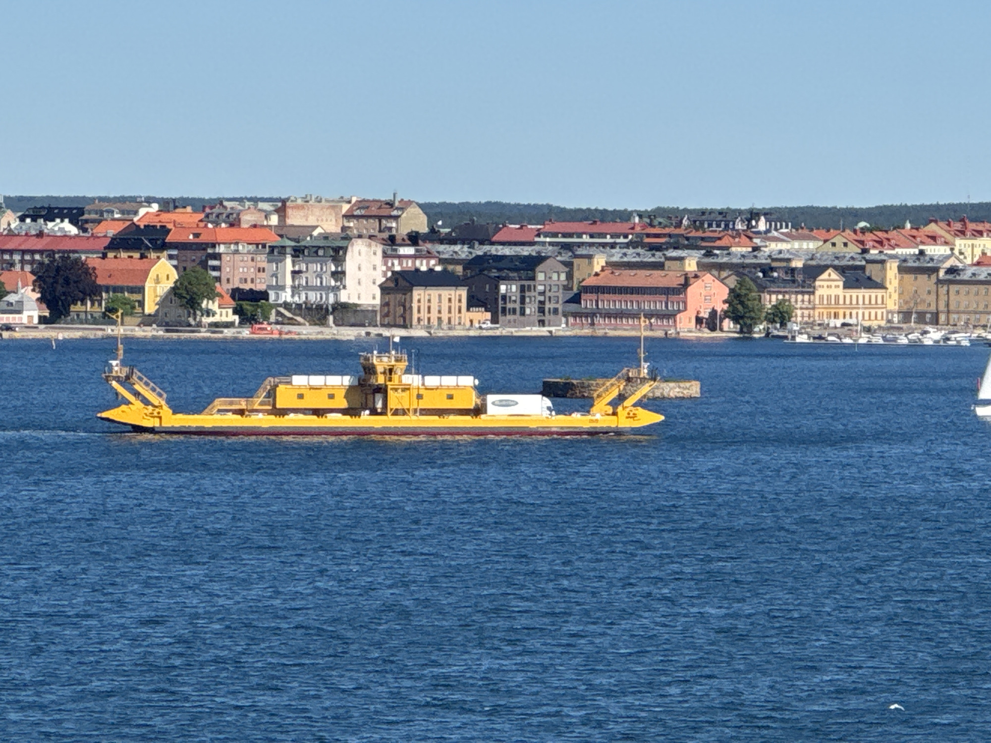 A yellow ferry boat, carrying a white truck, traverses the blue waters of a bay. In the background, the city of Karlskrona, Sweden, is visible. The city features a dense collection of buildings with red, orange, and yellow rooftops, interspersed with darker-colored structures and some green trees. The sky is a clear, bright blue, with no clouds present. The ferry appears to be moving from left to right in the frame. The water is choppy with small waves.
