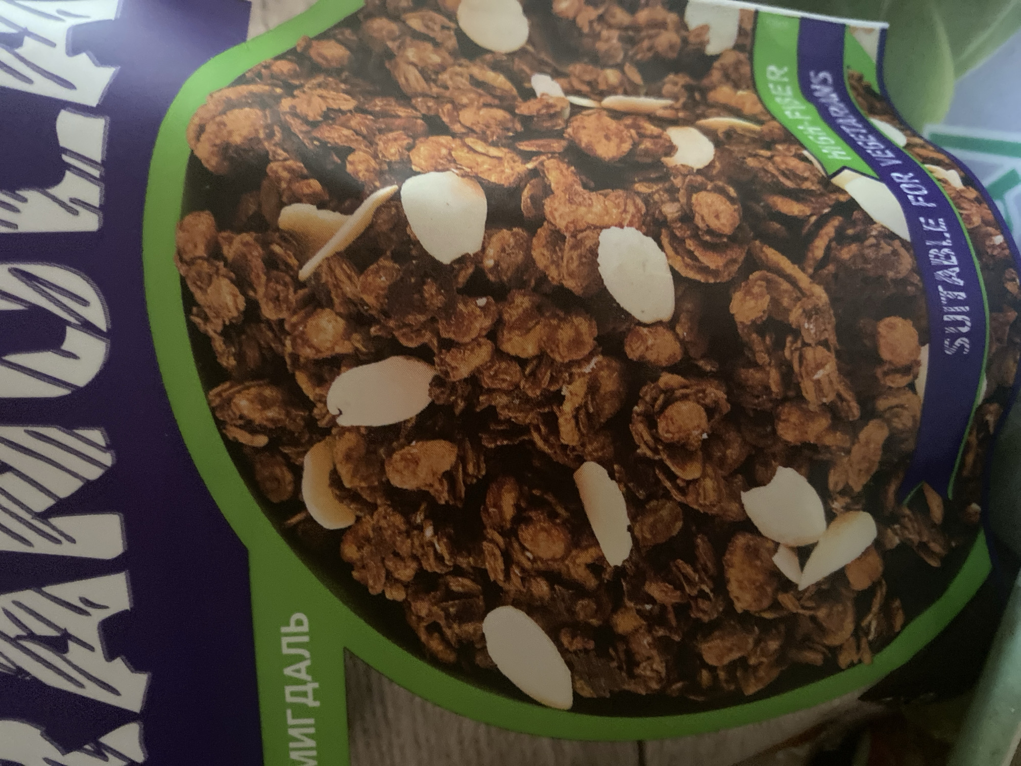 The image is a close-up shot of a bag of granola. The granola itself is a mixture of oats, nuts, and possibly chocolate, with visible slivers of almonds scattered throughout. The packaging is primarily purple with a green border framing the granola. Text is visible on the packaging, with "МИГДАЛЬ" in green text on the left side, which translates to "almond" in Russian. On the bottom right, a ribbon-like design with text reads "HIGH-FIBER" in green and "SUITABLE FOR VEGETARIANS" in white. The background is blurred and appears to be a textured surface, possibly wood. The overall lighting is consistent, suggesting an indoor setting. No people or specific locations within a city are depicted.