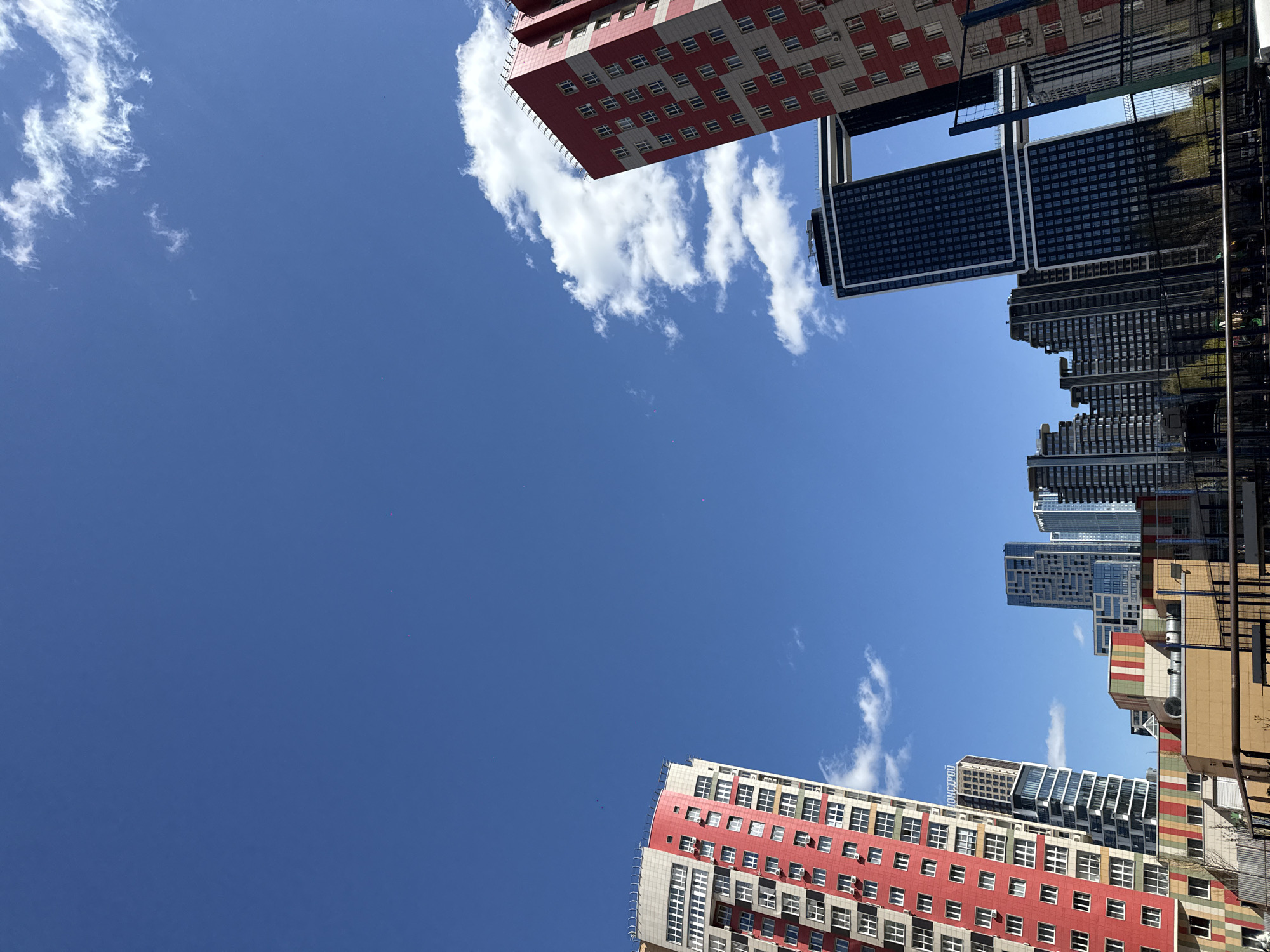 The image shows a low-angle view of a sky in Khimki, Russia, framed by buildings. The sky is a clear, deep blue with scattered white, puffy clouds near the upper left and lower right corners. Buildings are visible in the upper and lower portions of the frame. The buildings are multi-story structures. The color palette of the buildings ranges from red and white to dark grey and beige. Some buildings have a checkered pattern on their facades.