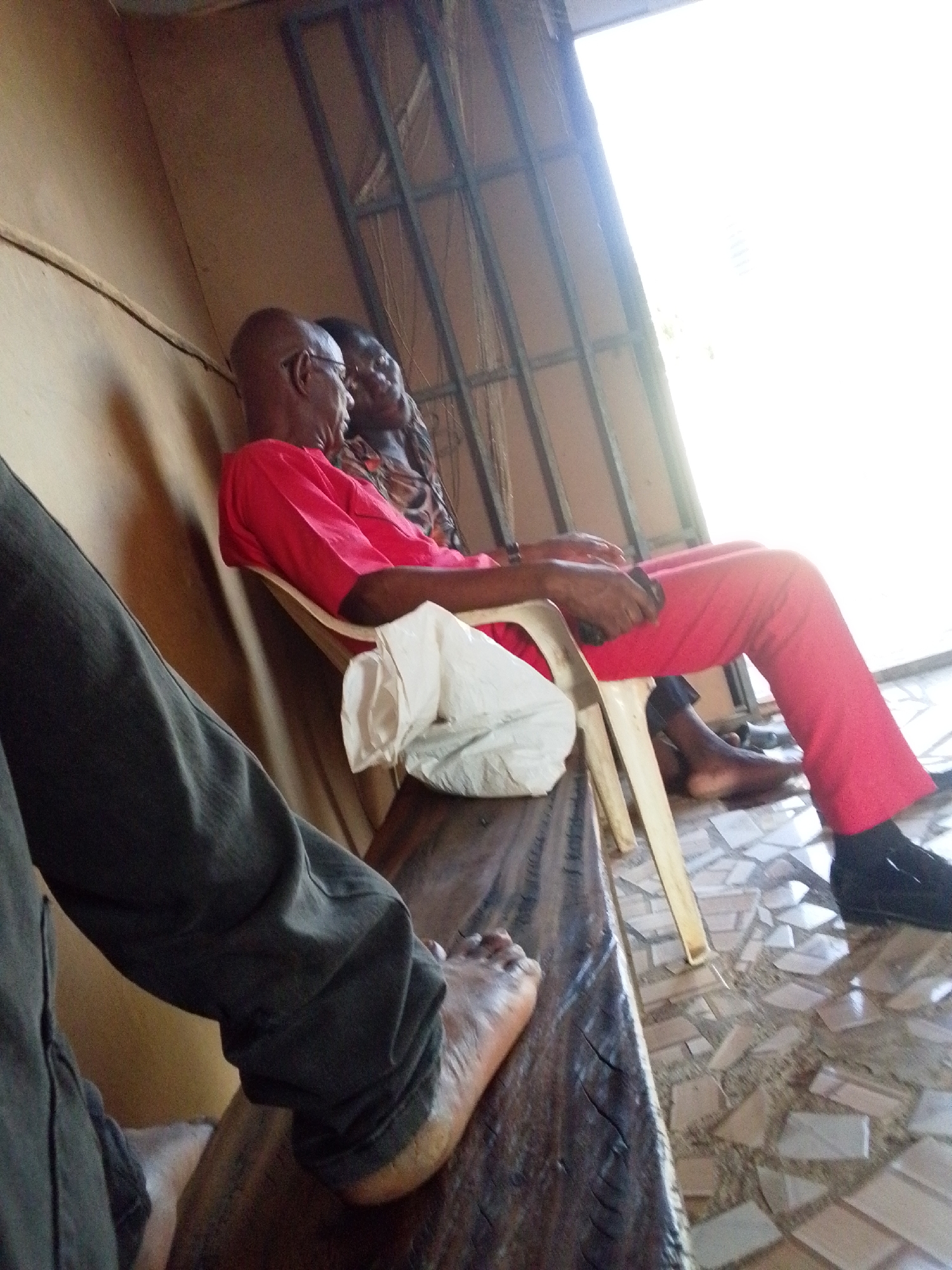 The image shows two older individuals seated in an indoor space. A bald man wearing glasses and a red shirt sits on a white plastic chair, with a white plastic bag hanging from the chair. A woman is seated next to him; her clothing is patterned. The man wears black dress shoes. The floor is tiled with a mosaic pattern. In the foreground, the lower part of a person wearing dark clothing is visible, with a bare foot resting on a dark wooden surface. The location is presumed to be Lokoja, Nigeria. A window provides bright light, casting shadows in the room.