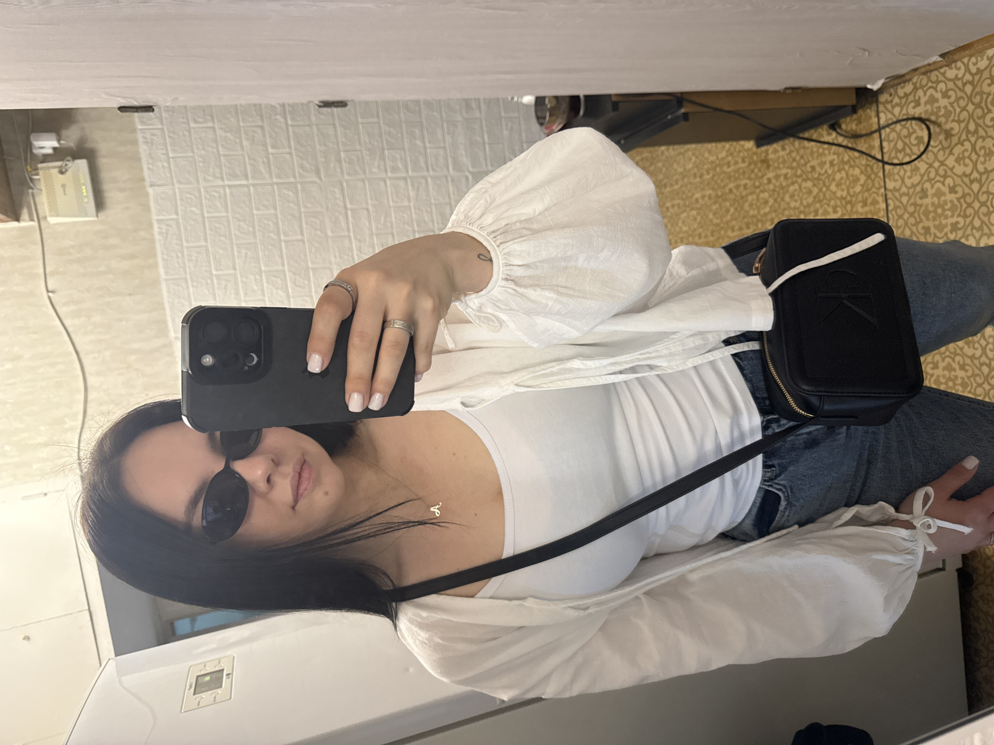 A woman takes a selfie indoors, presumably in her home. She is wearing sunglasses, a white tank top, a white blouse with puffy sleeves, and blue jeans. She also has a black Calvin Klein crossbody bag. The background includes a wall with textured panels and a glimpse of a floor with a patterned rug. There is no direct evidence to place this photo specifically in Mytishchi, Russia.