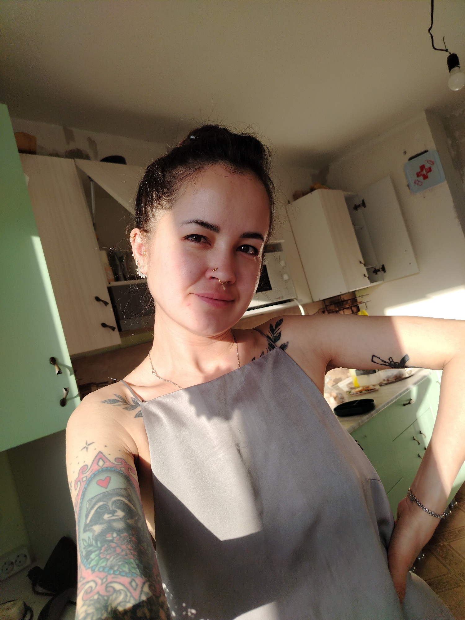 A woman with dark hair pulled into a bun is standing in a kitchen. She is wearing a gray garment, possibly an apron or dress. She has multiple tattoos on her arms, including one of a raccoon in a frame and another of leaves. She also has piercings in her ears and nose. The kitchen has green and white cabinets, some of which are open. A microwave is visible on the countertop. The wall shows signs of renovation, with unfinished plaster. A light fixture hangs from the ceiling, and a small decorative item with a red cross is attached to the wall. The location is Noviy, Russia.