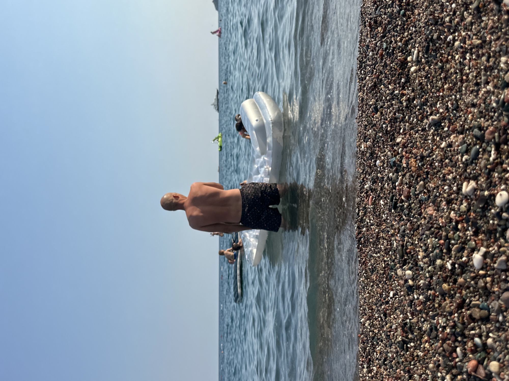 This image depicts a sunny day at a pebble beach in Boreti, Montenegro. In the foreground, the dark, multi-colored pebbles of the beach are visible, with the gentle waves of the clear blue sea lapping at the shore.
In the water, a man with a bald head is standing, facing away from the camera, wearing dark patterned swim shorts. He is waist-deep in the water. In front of him, a white inflatable raft is floating, and a person is partially visible on it, leaning forward. Further out in the sea, another inflatable raft with a child sitting on it can be seen. In the distance, a few other people are visible on inflatables or swimming, and a large white yacht is anchored. The sky is clear and blue.