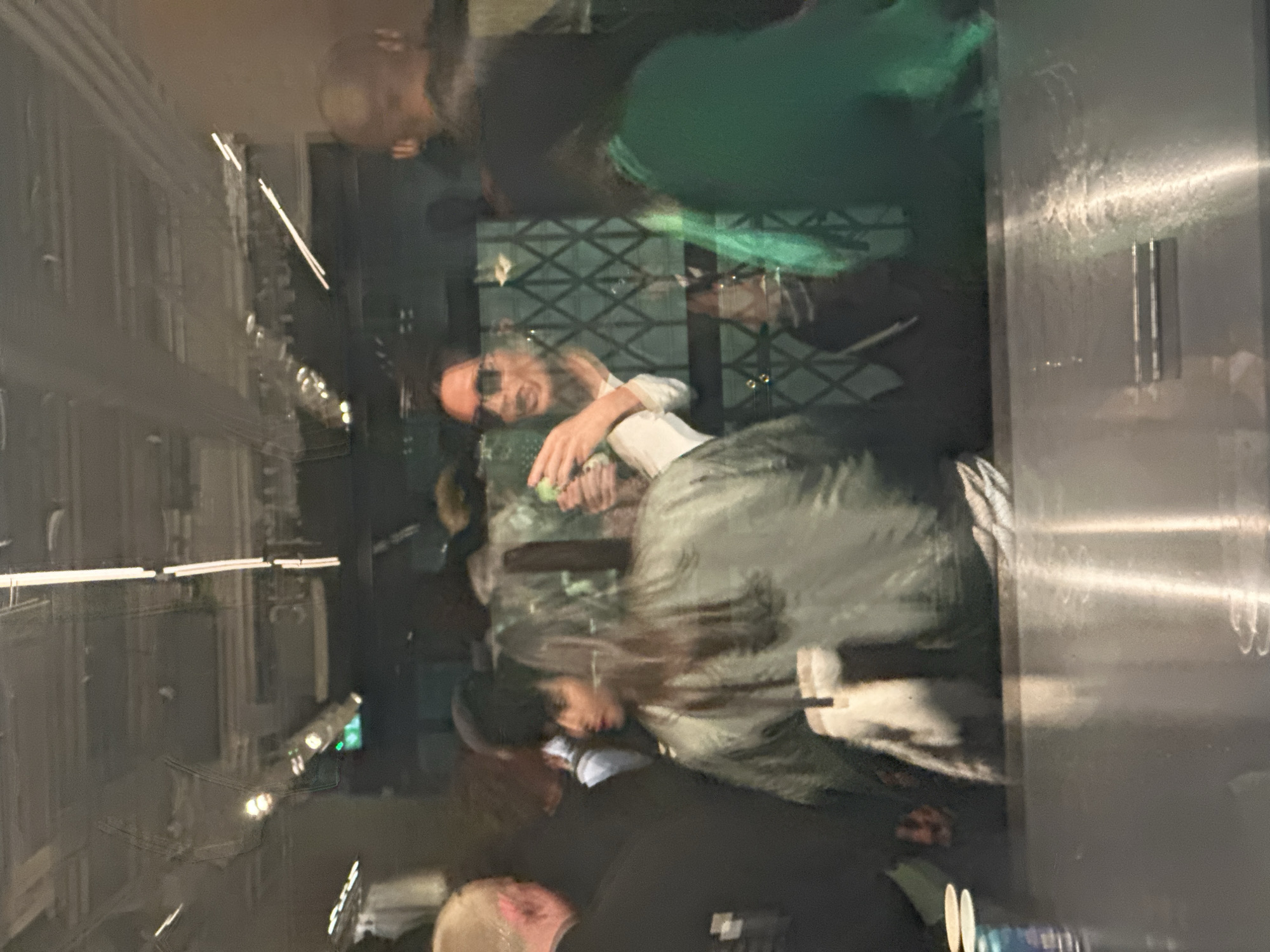 This medium-angle indoor shot captures a dynamic scene primarily through a reflective surface, likely a window or glass panel, creating a layered and somewhat abstract view. The image is oriented with the longer side horizontal.
Several individuals are visible, often blurred or as reflections. In the central-left area, a person with light-colored hair and sunglasses is prominent, wearing a white collared shirt under a dark jacket. This individual appears to be smiling or laughing, holding a small green object or drink. Behind them, a dark, patterned screen or grate is discernible.
To the upper right of this central figure, another person's head with short, light hair is visible. Further down and to the right, a person with long, dark hair is seen, partially obscured. In the bottom-right corner, another blurry head is present, along with a faint reflection of what might be a bottle or container.
The scene is dominated by reflections of artificial lighting, appearing as bright horizontal and vertical lines, suggesting an evening or nighttime setting. The ambient light indicates a dim, indoor environment, possibly a bar, restaurant, or lounge. A shiny, vertical metallic surface is visible along the far right edge of the frame, also showing reflections. The overall impression is one of social gathering and interaction, viewed in a complex, multi-layered visual field. No legible text is visible.