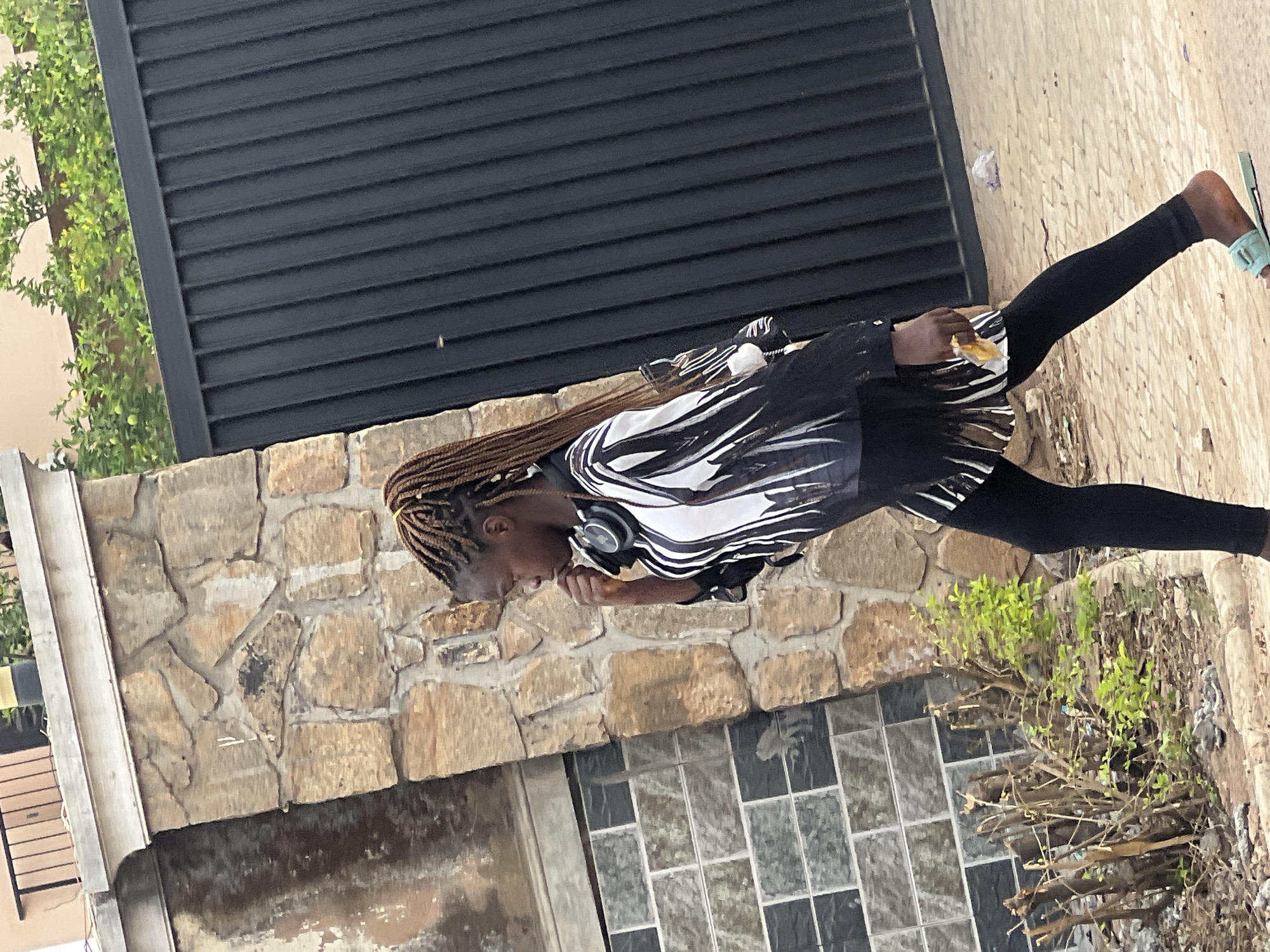 An outdoor, eye-level, full-shot image, presented rotated 90 degrees clockwise, captures a young woman walking in what appears to be an urban or suburban setting in Gura Top, Nigeria.
The woman has light brown braided hair and is wearing a black and white patterned long-sleeved top with black leggings and light blue or mint green flat sandals. A pair of black over-ear headphones rests around her neck. She is actively eating a snack, which appears to be a piece of bread or pastry, holding it up to her mouth with her right hand and looking down at it.
The background features a dark, horizontally slatted fence or gate to the left. Below this, a rustic stone wall made of irregularly shaped, light brown and tan stones forms a boundary. The ground she is walking on transitions from a patterned, possibly tiled or paved surface to an unpaved area containing dry brush and sparse green foliage. More vibrant green vegetation is visible beyond the fence on the far left.
The lighting suggests it is daytime with bright and clear weather conditions. Her expression is focused on her snack, and no specific emotional state is clearly discernible. There is no visible text in the image.