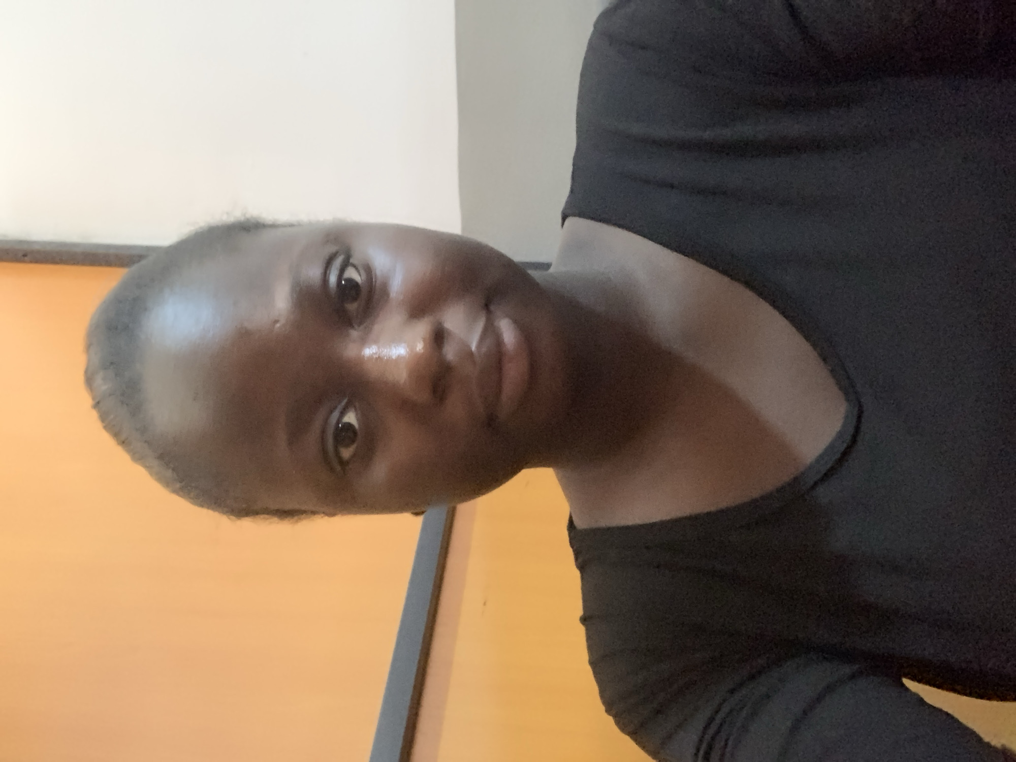 A medium shot captures a young Black woman from the chest up, facing forward in a head-on selfie. She has dark skin and her hair is pulled back tightly, revealing a prominent forehead with a slight sheen from oil. Her eyes are dark and wide open, looking directly at the camera. A faint mole is visible on her left cheek. She wears a simple black t-shirt with a V-neckline. The background consists of an orange wall on the left and a lighter, possibly white or beige wall on the right, separated by a dark horizontal line. The lighting appears to be overhead, creating some highlights on her skin. The scene suggests an indoor setting, possibly a room or an office.