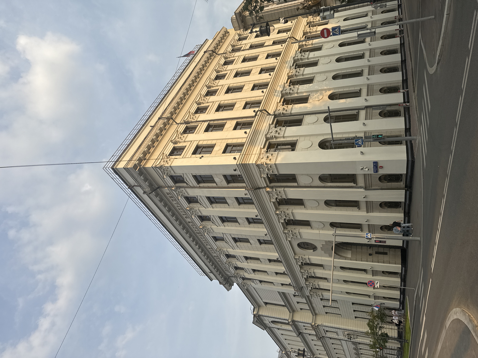 The image depicts a wide-angle, eye-level view of the Supreme Court of the Russian Federation building in Moscow, Russia, captured on a clear day with scattered clouds. The building, a grand neoclassical structure, is predominantly white with numerous windows and ornate columns. The sun casts shadows on the facade, highlighting the architectural details.
In the foreground, a paved street with crosswalks and traffic signals is visible. Two pedestrians are standing near a traffic signal and a no-parking sign. Further down the street, a group of four people are walking on the sidewalk next to some trees.
Above the building, a power line stretches across the sky. A Russian flag is subtly visible at the top of the building. The overall scene is a snapshot of urban life in Moscow, with the imposing architecture of the Supreme Court as the central subject.