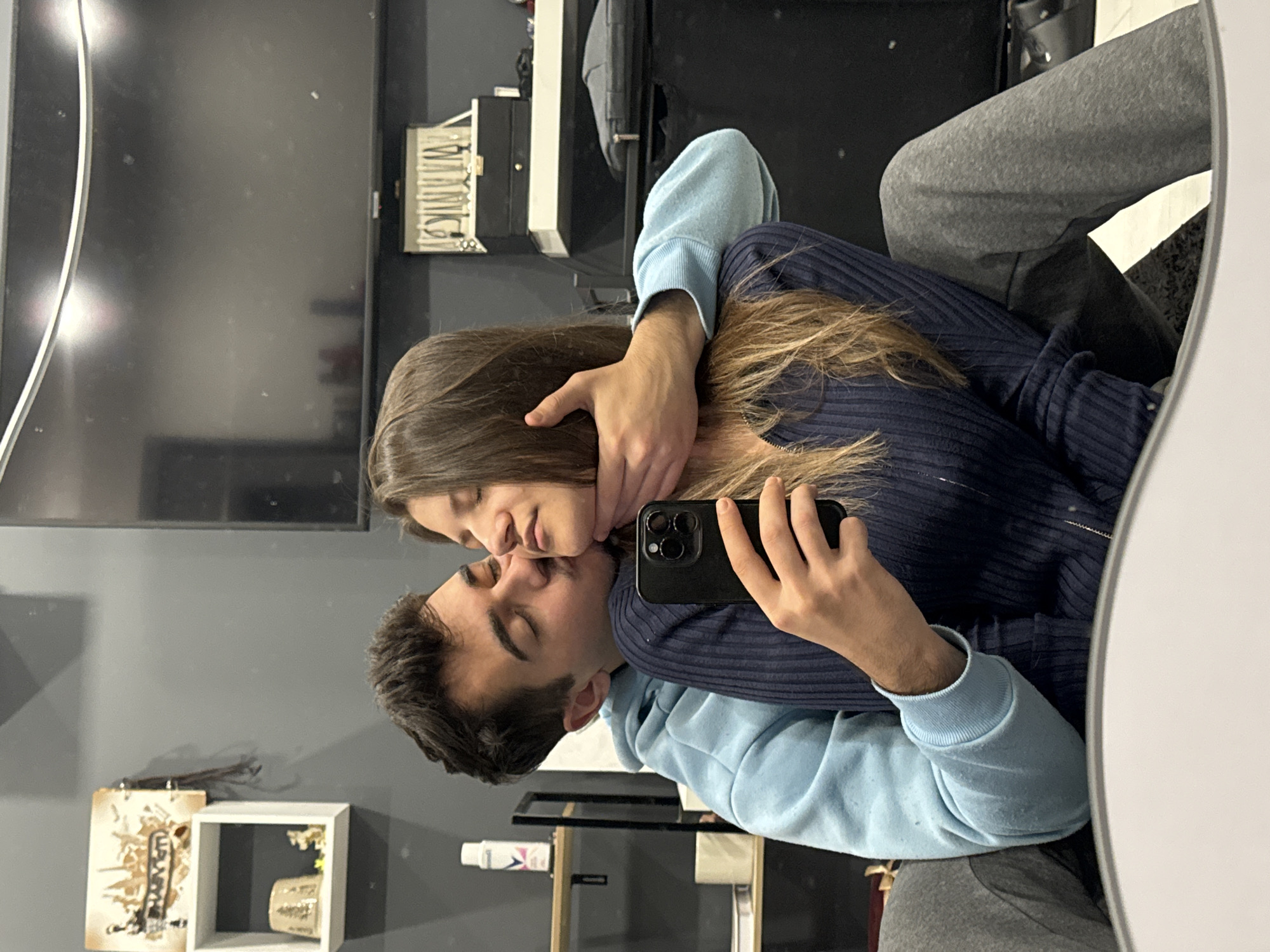 A medium shot taken at eye level depicts a man and a woman kissing. The man has dark hair and is wearing a light blue hoodie. The woman has long brown hair and is wearing a dark blue ribbed sweater. The man is holding a black smartphone. The background includes a grey wall, a television, and a shelf with decorative items. The image is likely taken in an apartment. There is no information available to confirm if this image was taken in Sofia, Bulgaria.
