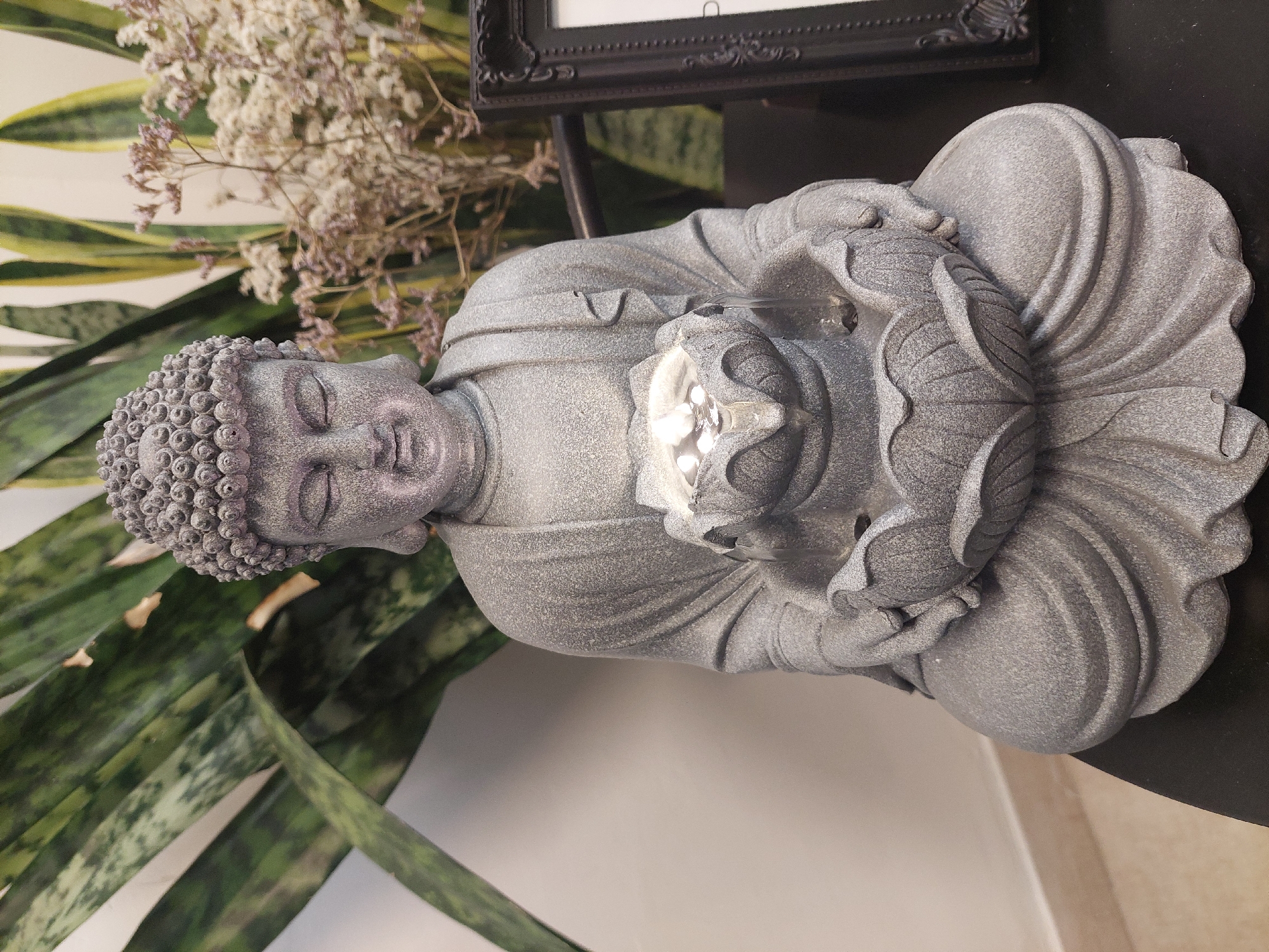 The image captures an indoor decorative display featuring a gray, textured Buddha statue in a meditative posture. The statue is depicted seated with its hands clasped, holding a lotus-shaped object that emits a bright, cool light from its center. The Buddha's face conveys a serene expression with closed eyes, and its head is adorned with characteristic tightly curled hair. The textured surface of the statue suggests it is made of stone or a stone-like material.
To the left and in the background, vibrant green and yellow variegated leaves, likely from a snake plant (Sansevieria trifasciata), provide a natural backdrop. Behind the statue and plant, a cluster of dried, light-colored flowers or foliage adds a delicate, muted tone to the composition. A portion of an ornate black picture frame is visible in the upper right background, resting on what appears to be a dark shelf or table, on which the Buddha statue is also placed. The background consists of a plain, light-colored wall.
The scene is well-lit, likely by ambient indoor lighting, accentuating the details and textures of the objects. No specific activity or event is taking place; the image is a static portrayal of a decorative arrangement. The overall aesthetic evokes a sense of peace and contemplation. There is no visible text in the image. This scene is located in Buenos Aires, Argentina.
