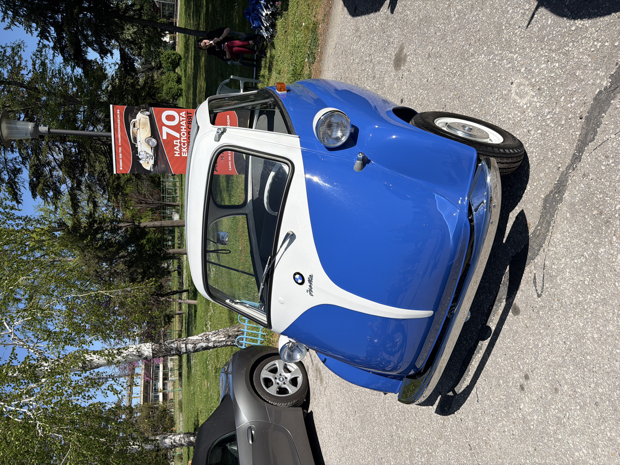 The image shows a blue and white BMW Isetta, a small car, parked on an asphalt surface, likely a street or parking area. A silver convertible car is partially visible next to it. A sign on a lamppost behind the Isetta advertises an exhibition with "70 над експоната" (70+ exhibits) and mentions "MARKOVOMUSEUM.BG", the website for a museum in Plovdiv. People are sitting on chairs in the background on a patch of grass. The location is Plovdiv, Bulgaria, based on the text on the sign.