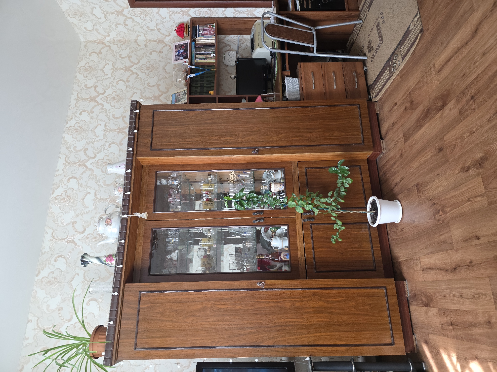 The image depicts the interior of a room in Qarabalıq, Kazakhstan. A large, wooden, floor-to-ceiling cabinet with glass doors dominates the left and central portions of the scene. This cabinet displays a collection of glassware and decorative items. To the right of the cabinet is a wooden desk with a computer monitor, printer, and a chair. The room has patterned wallpaper and a wooden floor. A plant in a white pot is positioned in front of the cabinet. The lighting suggests it is daytime, with sunlight casting shadows on the floor.