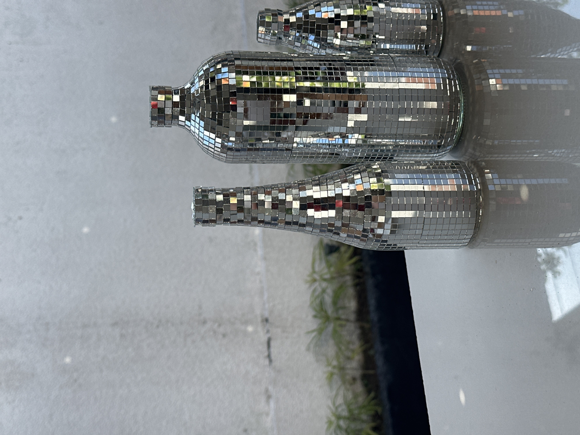 Three disco ball-covered bottles are arranged on a reflective surface. The bottles are of varying heights and widths. The largest bottle is in the center, a medium-sized bottle is to its left, and the smallest bottle is to the right of the largest. The background features a textured grey wall with a band of greenery below it. The scene is lit with bright light, creating reflections on the bottles and the surface they are placed on. There are no people or animals present in the image. The location is a still-life setup in North Kuta, Indonesia.