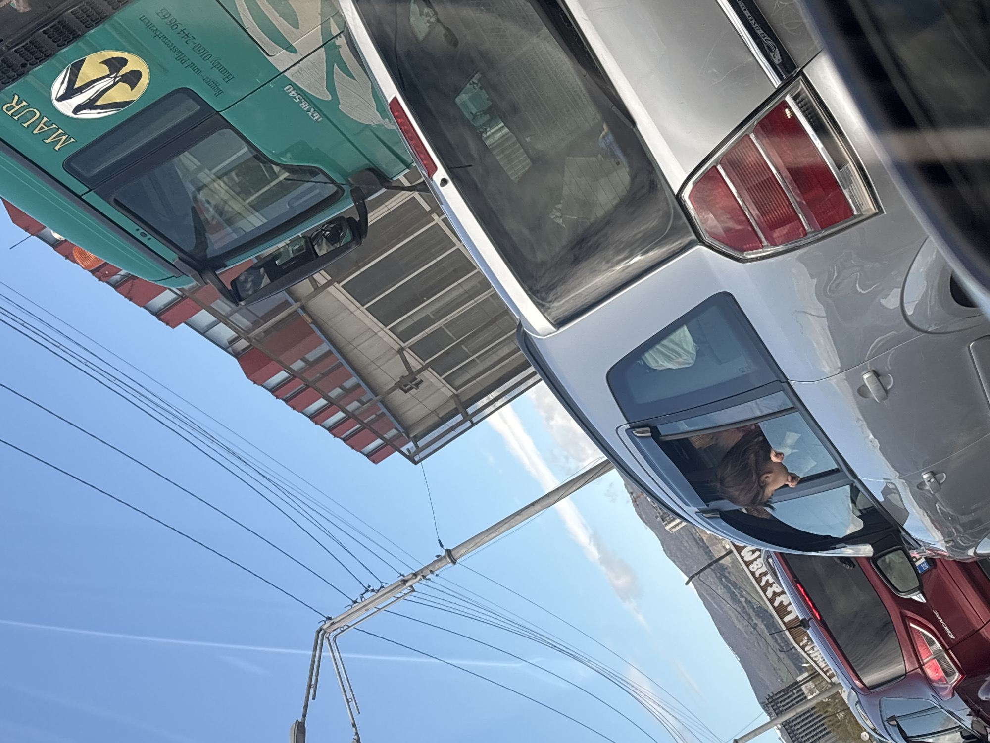 The image shows a street scene with several parked cars. The foremost car is silver with a sunroof through which a person's head is visible. To the left, a green truck with the word "MAUR" is seen, along with some contact information in German. Power lines stretch across the clear blue sky above the cars. Buildings can be seen in the background, and a glimpse of mountains is also visible. Based on the style of the buildings and vehicles, this location is likely in Tbilisi, Georgia.