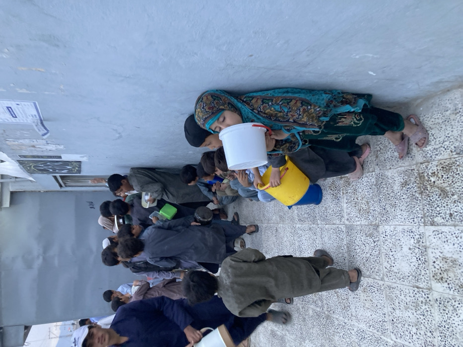 The image displays an outdoor gathering in Quetta City Tehsil, Pakistan, featuring approximately 25-30 individuals, predominantly children, positioned along a light blue-grey wall and an adjacent structure. Most individuals wear traditional shalwar kameez; several girls and women wear head coverings. In the immediate foreground, a young girl in a teal patterned dress and headscarf is drinking from a white plastic bucket held to her mouth, while also holding a yellow plastic bucket in her left hand. Another child next to her holds a blue bucket.
Numerous other children are either seated on the ground along the wall or standing in the open paved area, many holding various containers, including white plastic bowls, green plastic containers, and metal pitchers. A young male in a dark blue shalwar kameez and white cap, carrying a white metal pitcher, moves from the left side of the frame. A cluster of children is visible near an open doorway in the wall, above which several posters or papers are affixed. The ground is paved with light-colored, speckled tiles. A grey building or corrugated structure and faint distant mountains are visible in the background, suggesting an urban or semi-urban setting. The scene indicates a gathering, possibly for the distribution of resources.