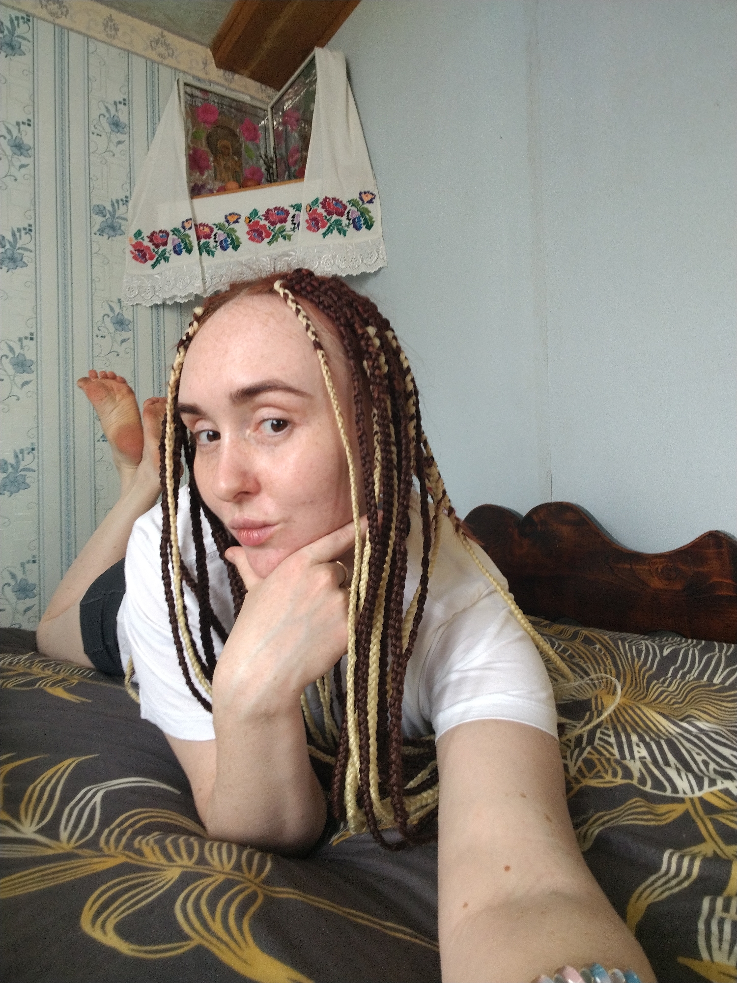 A fair-skinned woman with freckles and auburn hair styled in brown and blonde braids lies on a bed covered in a dark grey and gold patterned bedspread. She wears a white t-shirt and dark shorts. Her right arm is extended towards the camera, while her left hand supports her chin. Her feet are lifted behind her. The background includes a light blue wall, wallpaper with a floral pattern, and a framed religious image covered with a decorative towel. The location is likely a bedroom, but specific location details are unknown.